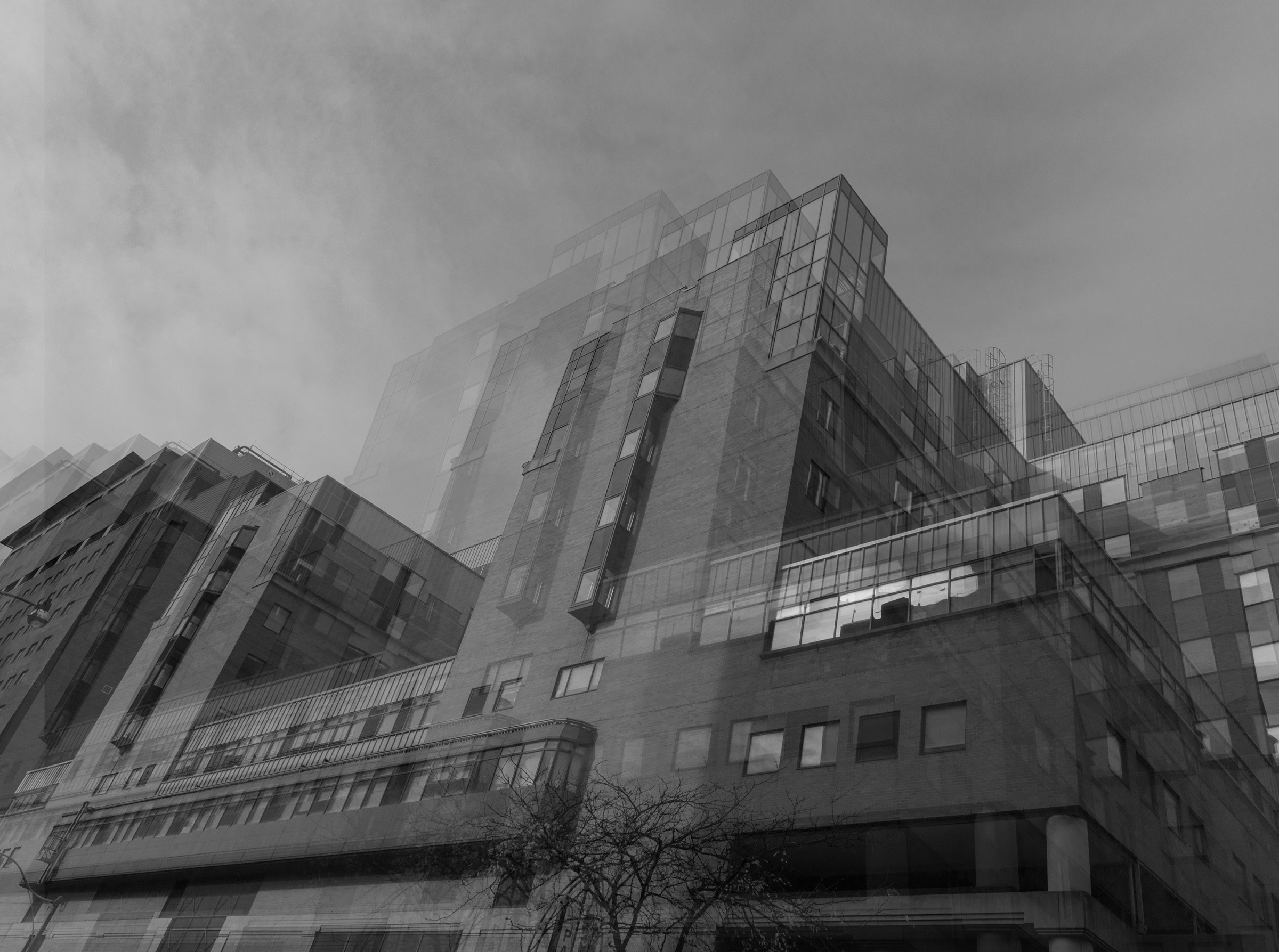 A black and white long exposure photo of the exterior of Sick Children's Hospital taken at the side of the hospital. There is a bare tree in front of the hospital, and a cloudy sky taking up almost half of the top of the photo.