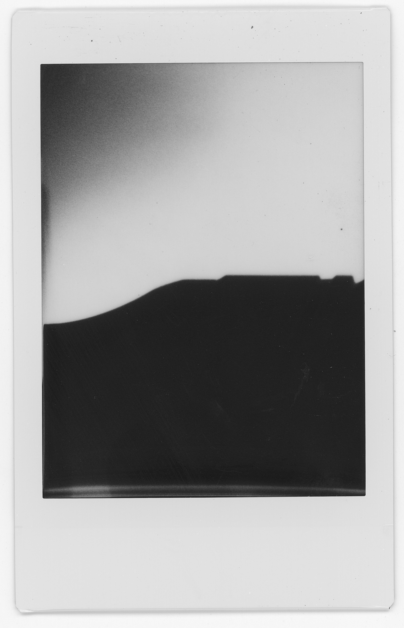 A black and white polaroid of a hill. The hill slopes up from the right of the image where it evens out half way through the image. The top of the photo above the hill is all white. There is a grey shadow in the top left corner.