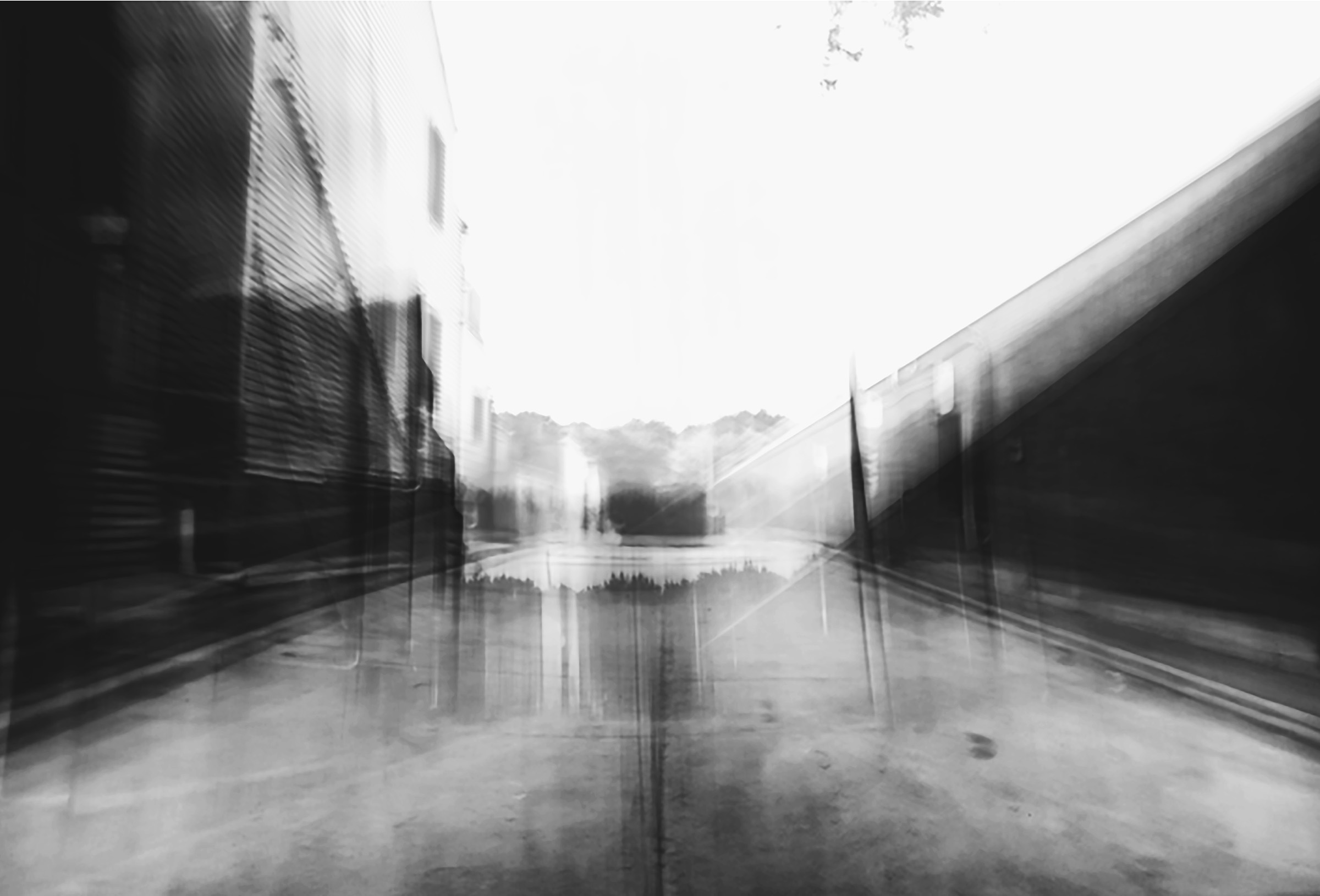 A black and white long exposure photo of an alley. There are sides of houses on the left of the alley and the back of a store on the right side of the alley. Behind the alley are trees.