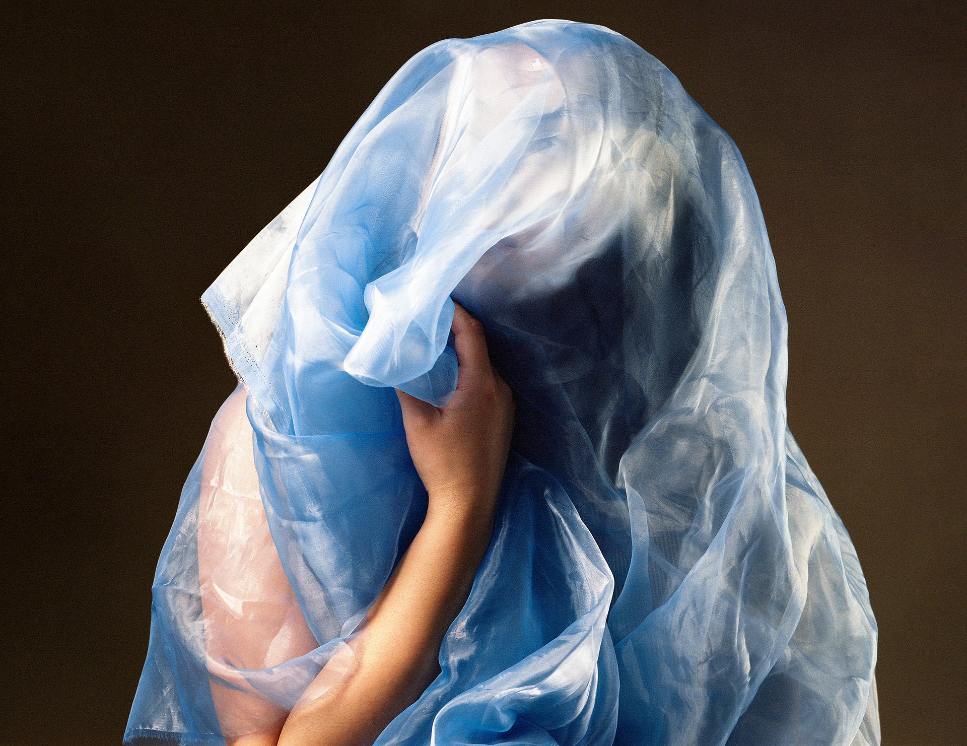 A photograph of someone wrapped in a blue sheer fabric, their hand grasps the fabric and their face is partially visible behind the fabric