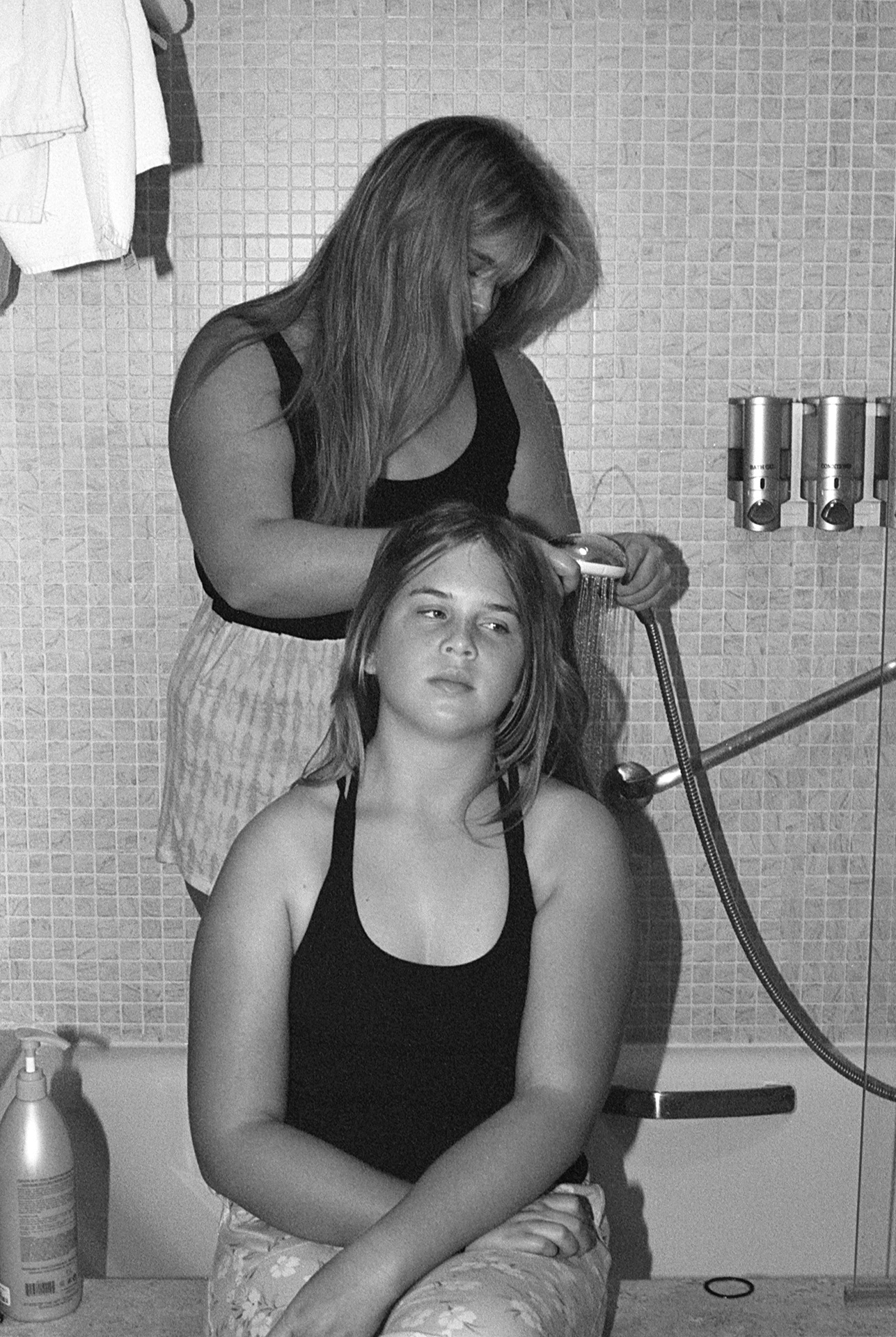 Eden sits on the edge of a bathtub, while Mariah stands inside of the bath with the shower head in hand. She is focussed on rinsing Eden’s hair, whilst Eden looks off into the distance.