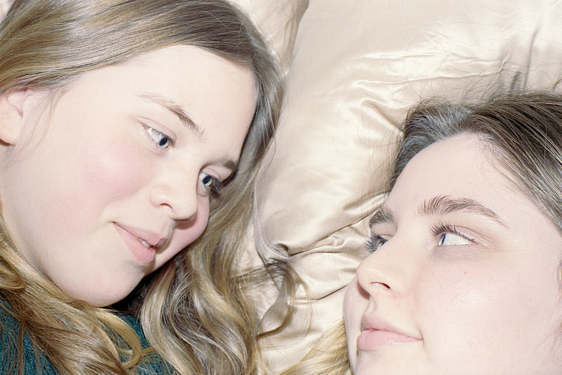 Mariah and Eden lay next to each other against a light pink silk pillowcase. They share direct eye contact while small smirks play on both of their mouths.