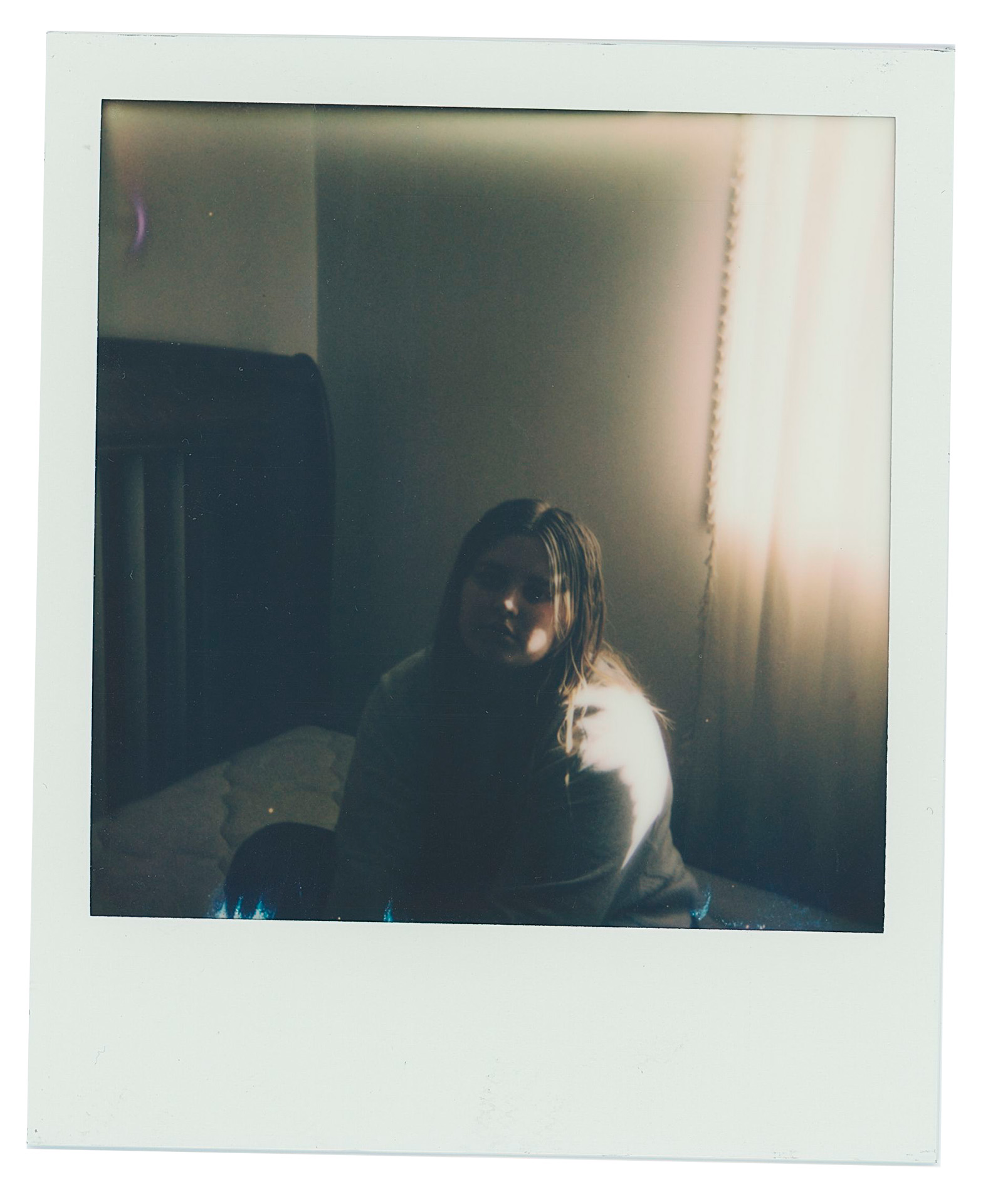 A Polaroid of Mariah sitting on a bed in her childhood room. The lighting is dim, with only a small amount of light coming from the window to her right.