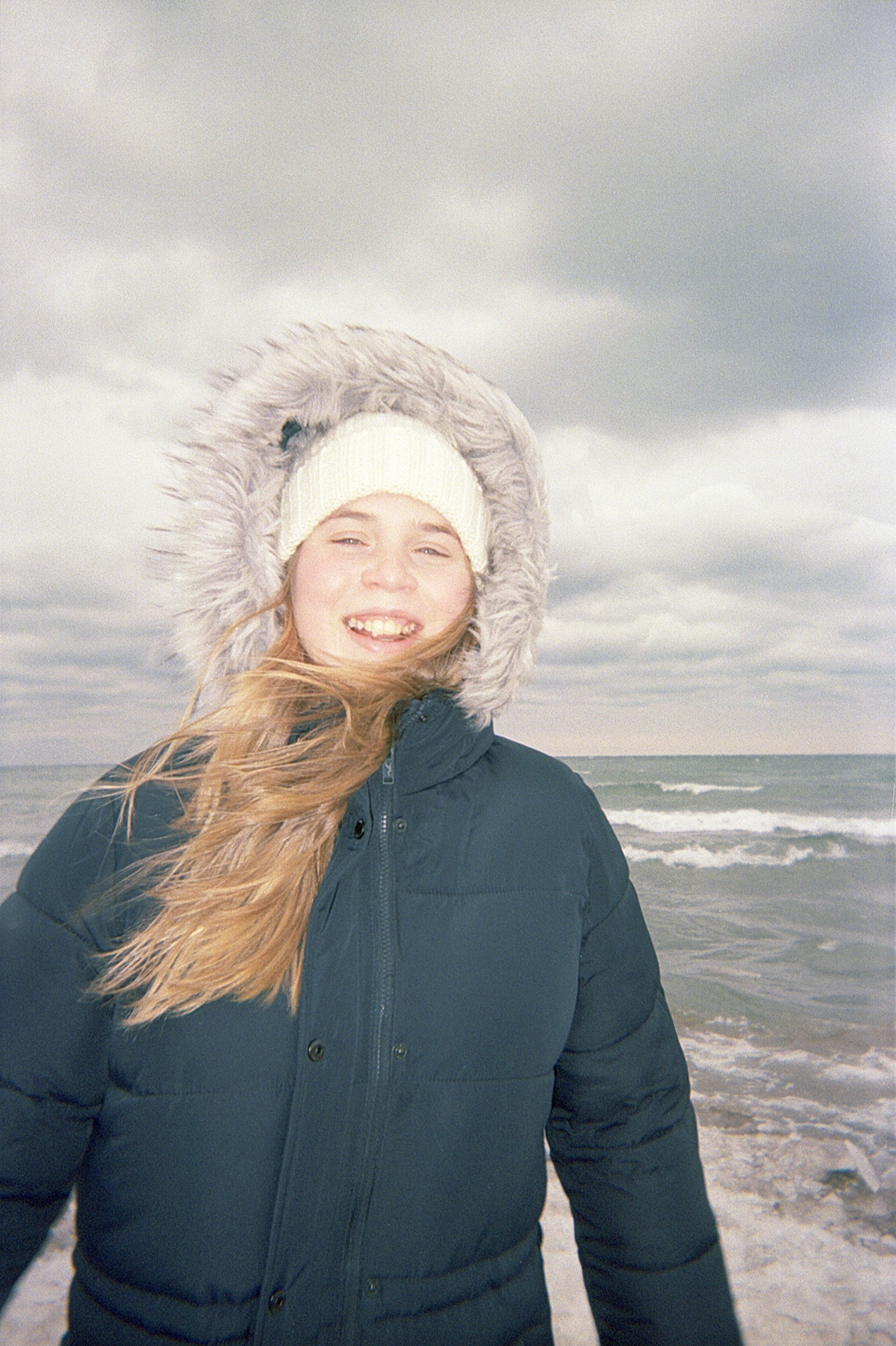 Eden laughing with her teeth showing. She is wearing her winter jacket with a fur hood pulled up. There are waves from Lake Ontario in the background.
