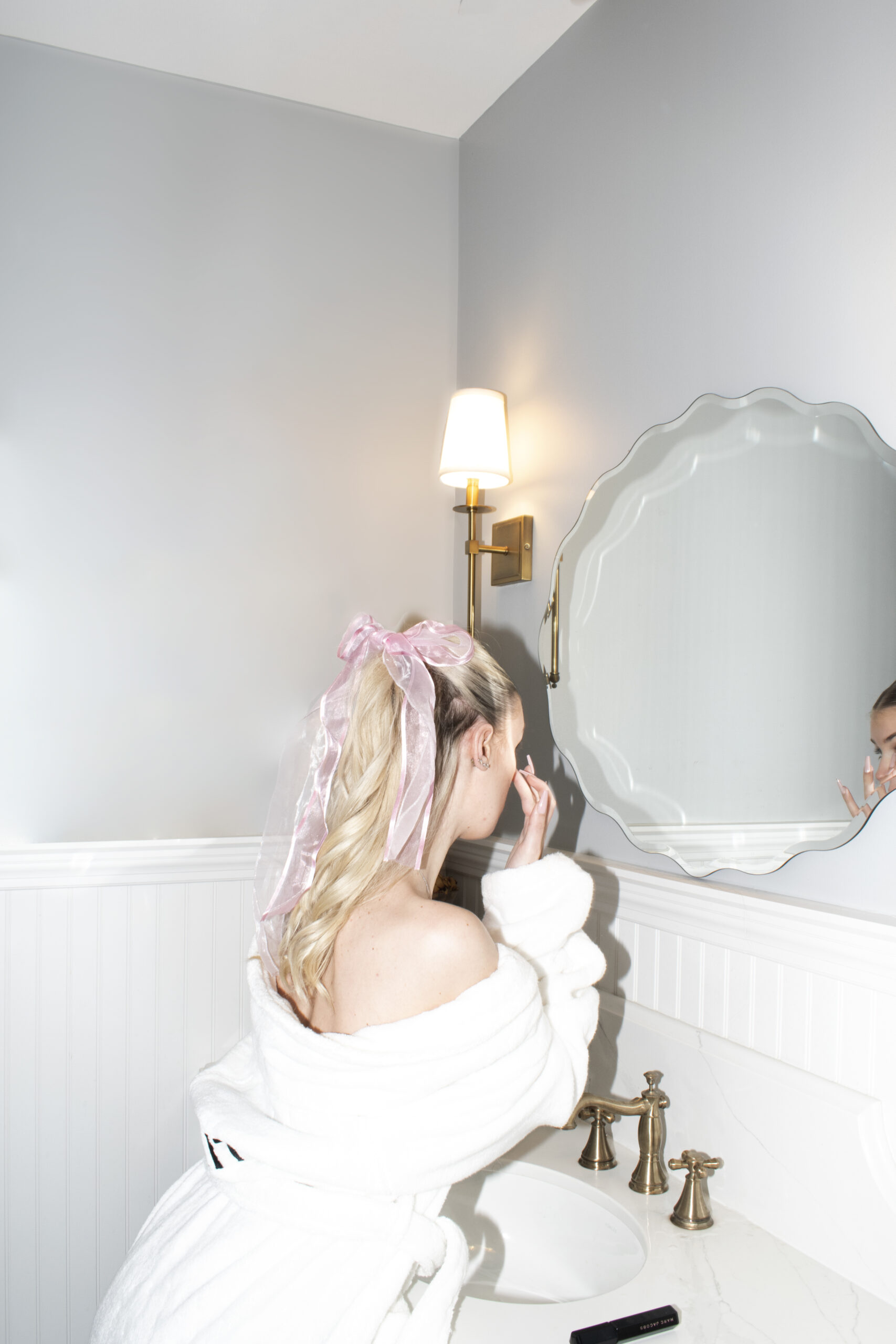 Photograph of a blonde woman wearing a white robe with a pink bow in her hair putting makeup on in front of the mirror.