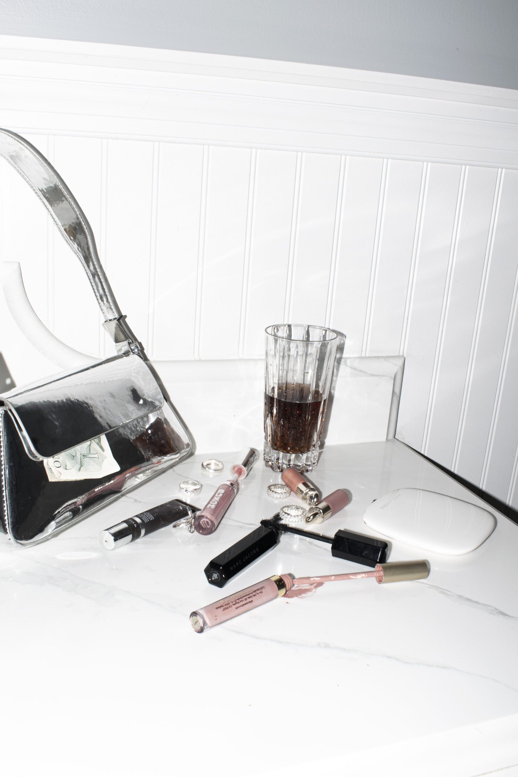A flay lay image of makeup items splattered onto a white countertop with a glass of wine and a silver metallic purse on the left.