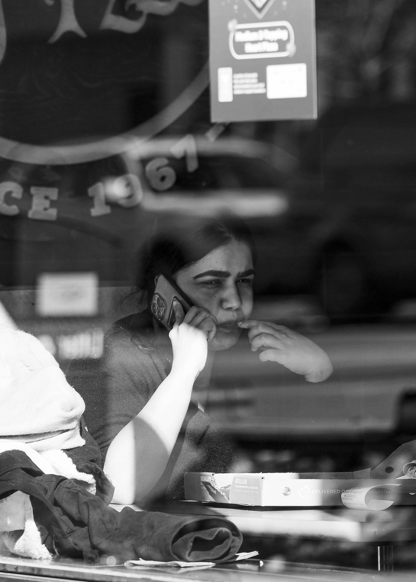 black and white image of a woman on a call while grabbing food shot through a window