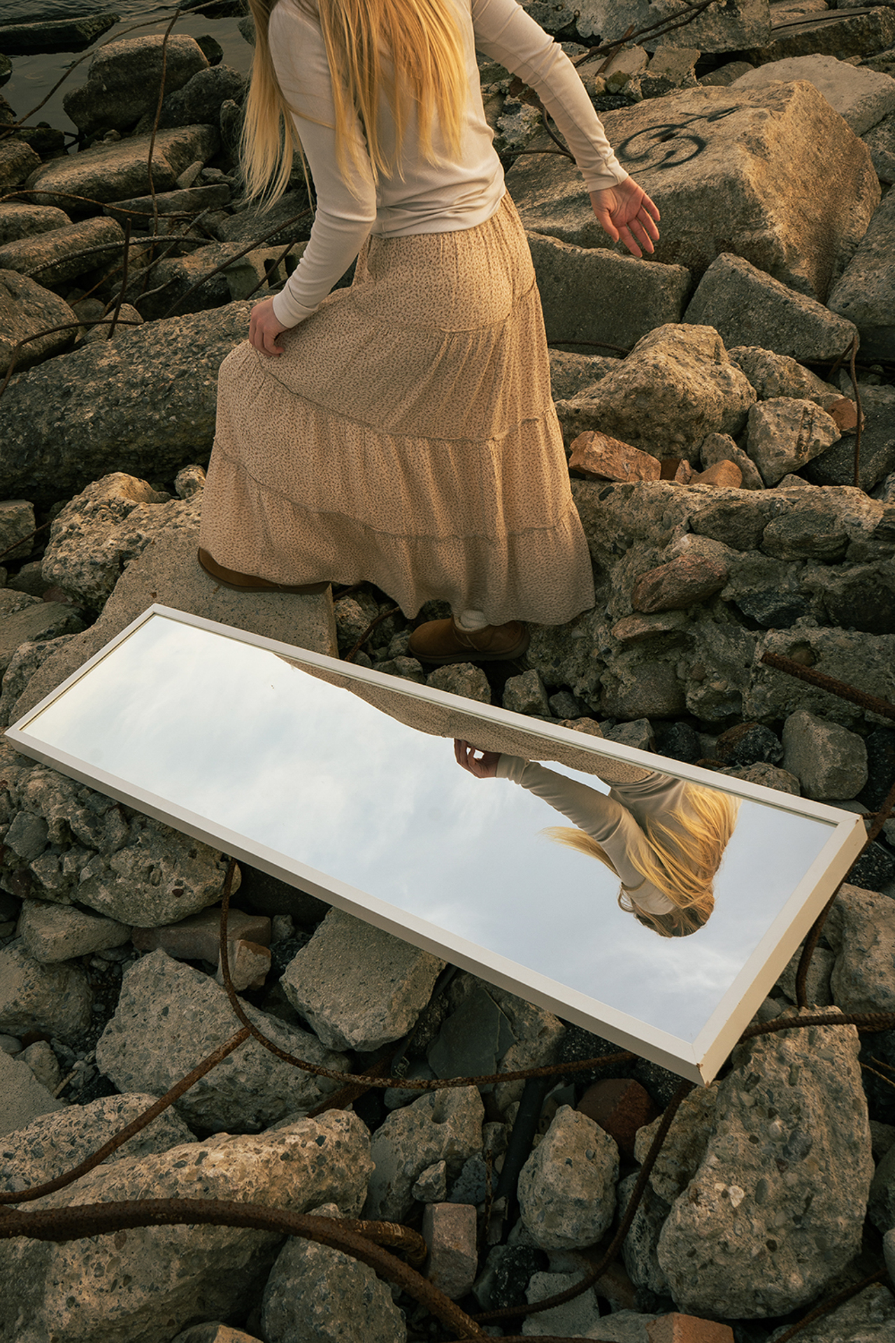 This photo consists of a mirror placed on the ground in a rock-field as a woman with blond hair walks along the rocks beside the mirror.