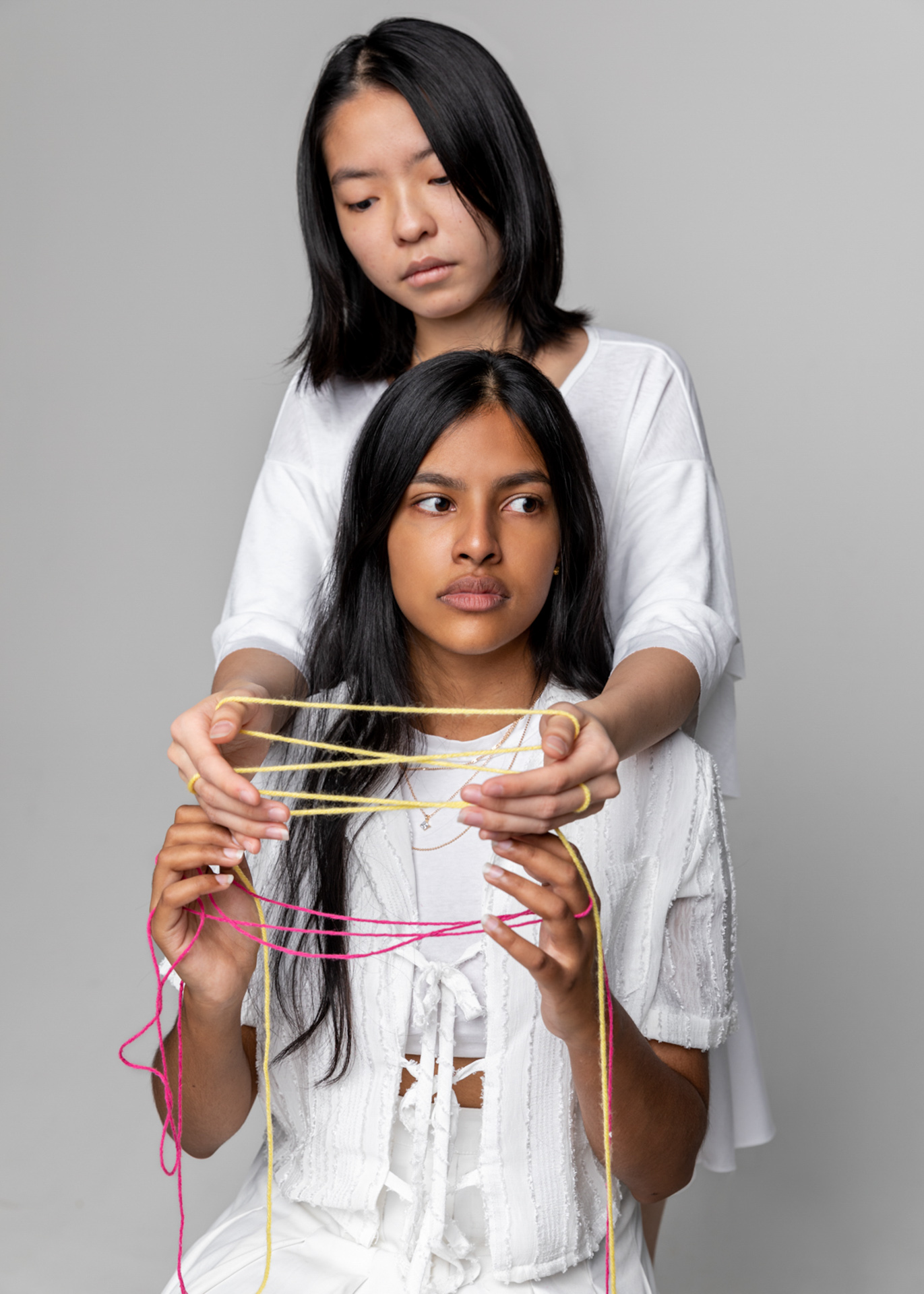 Two women, one sitting the other standing with her arms over the shoulders of the other. Their hands are intertwined with yarn looking away and at their hands. Both women are wearing white and the yarn is both yellow and pink.