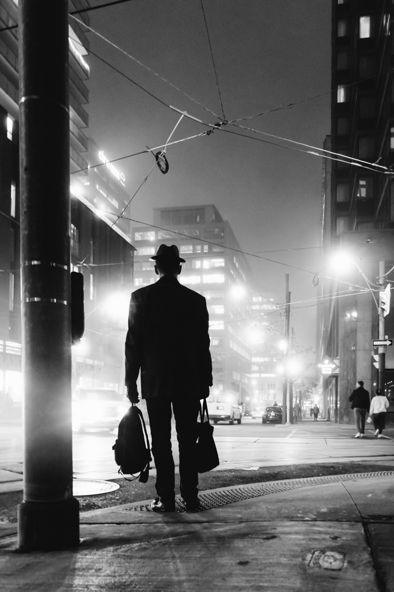 The photo depicted is in black and white and is taken of a man in a dress coat facing away from the camera holding a backpack in one hand and a tote bag in the other. He is standing at the corner of a street as bright streetlights fill the photo.