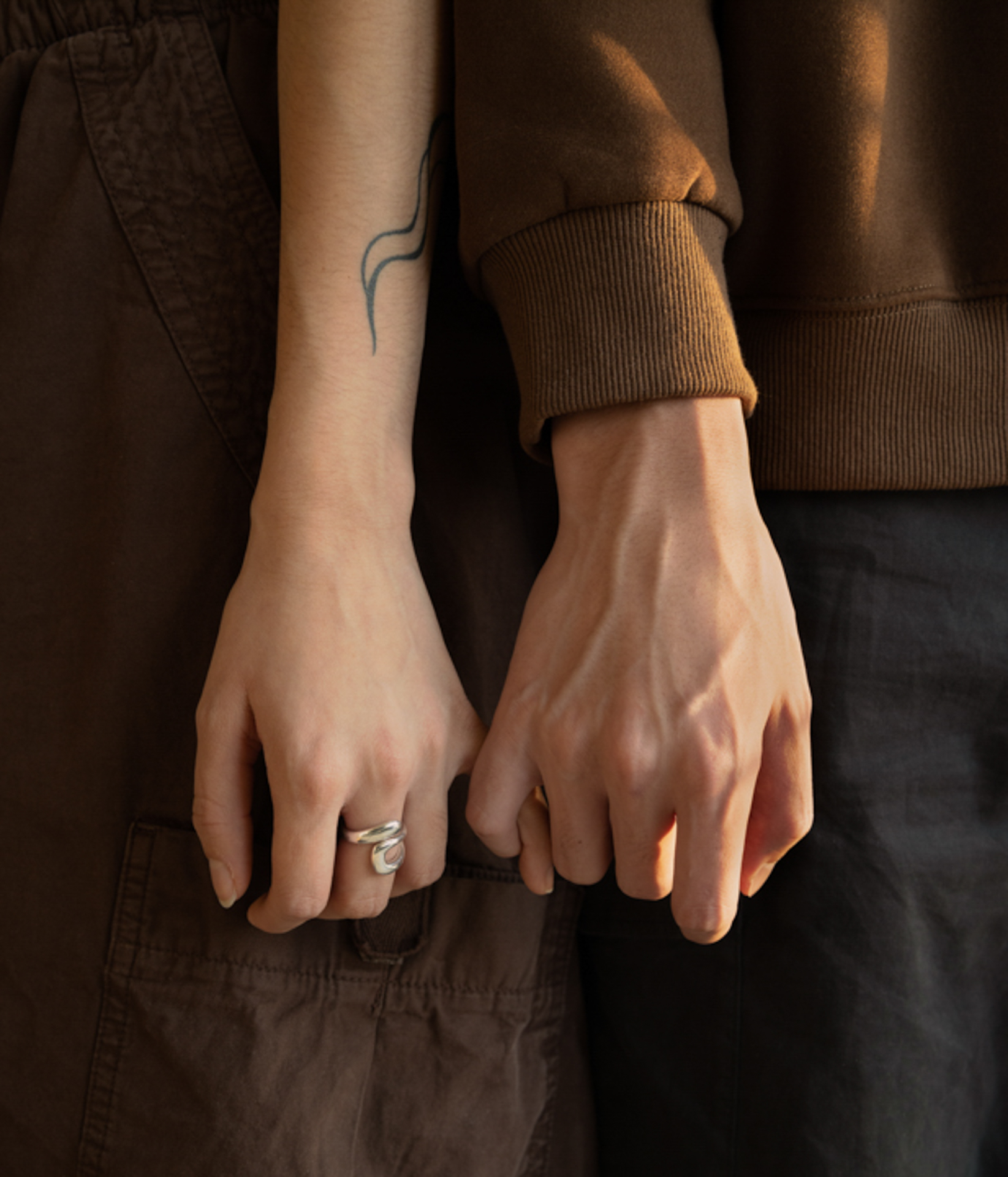 This photograph is a close up detail shot of two people's hands linked by their pinkies. The hand to the left is wearing a silver ring on their middle finger. The hands are at waist level and the overall colour scheme consists of warm browns and greys.