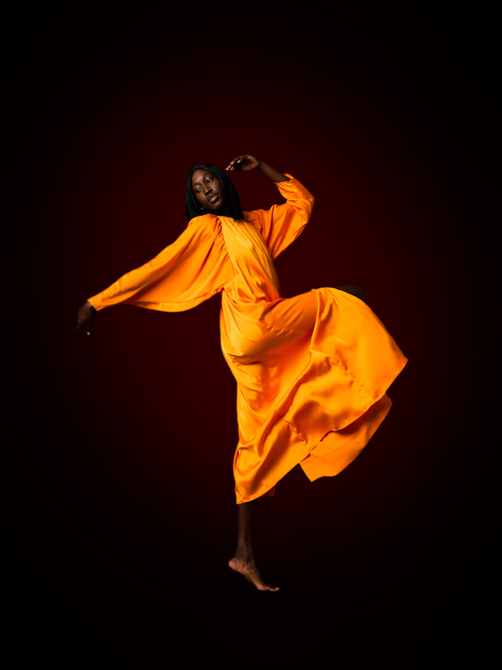 Black model in an orange silk dress kicking her leg and arms in the air against a dark backdrop.
