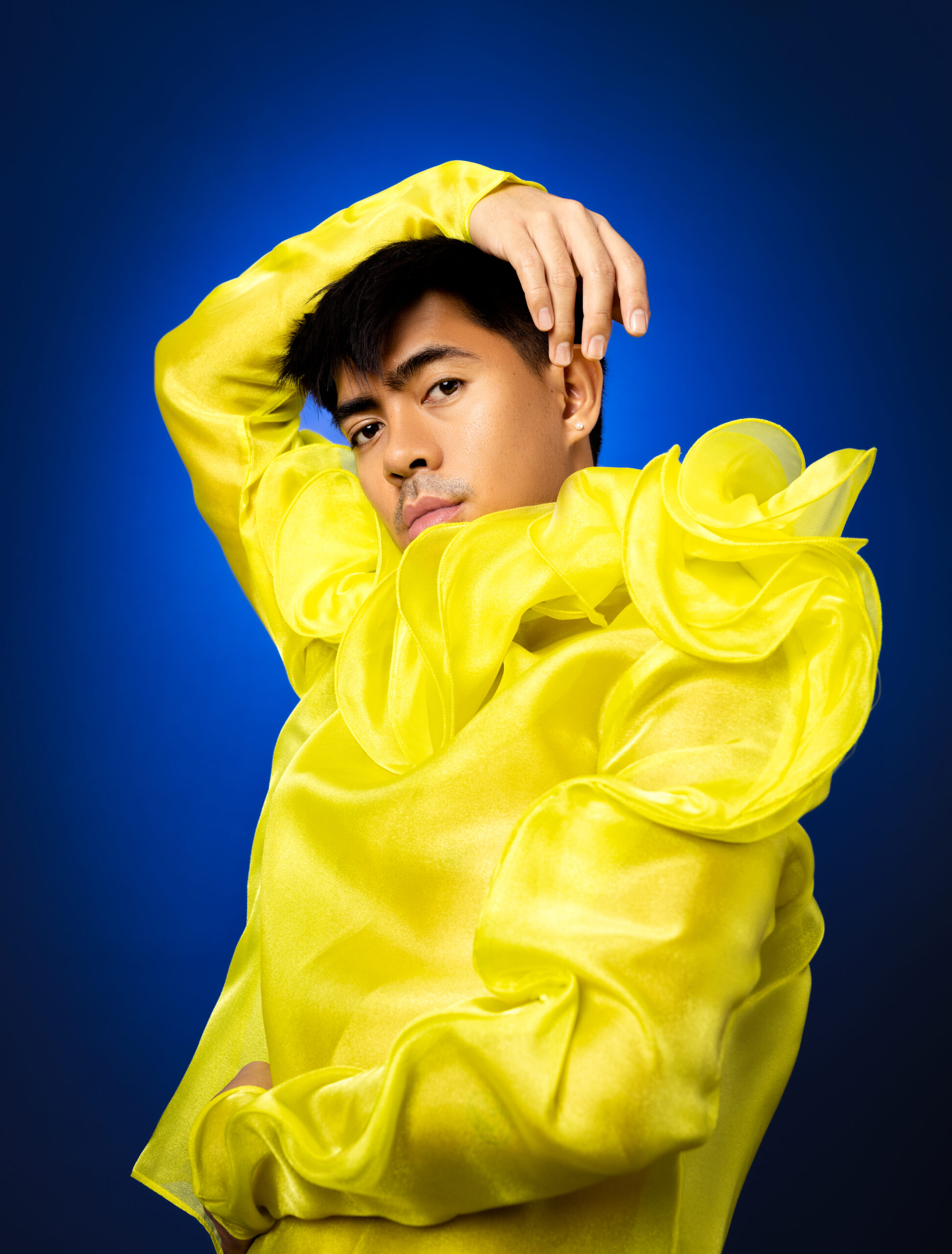 Asian model posing with one arm above head in a bright yellow top against a blue backdrop.