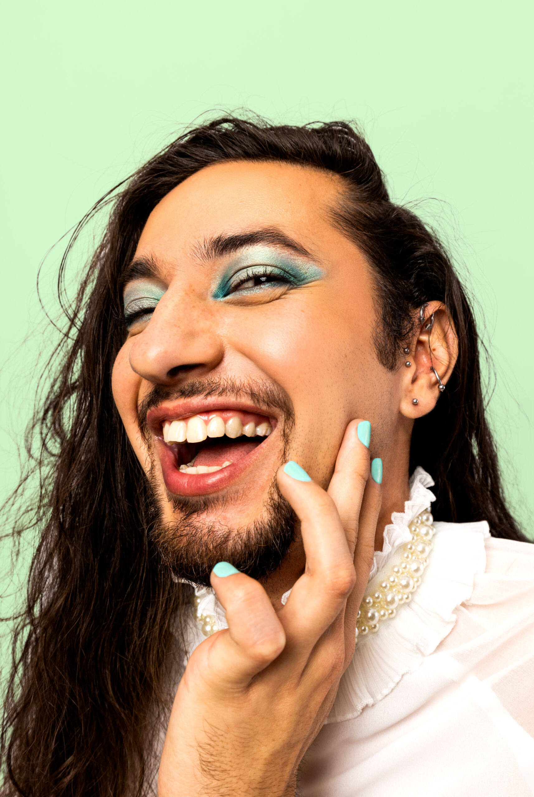 Indigenous model wearing blue eyeshadow and teal nail polish posing with hair against the face while laughing against a green backdrop.