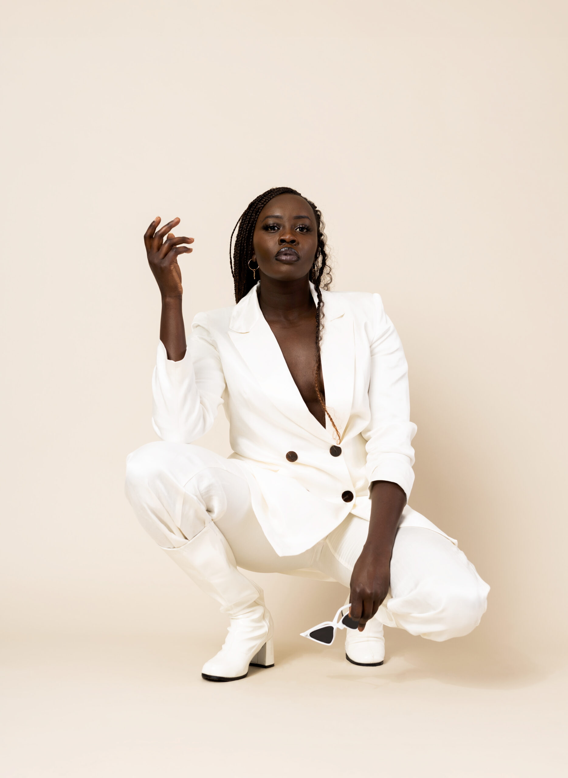 Black model crouching down while holding one hand in the air and the other hand holding sunglasses while wearing a white suit against a cream backdrop.