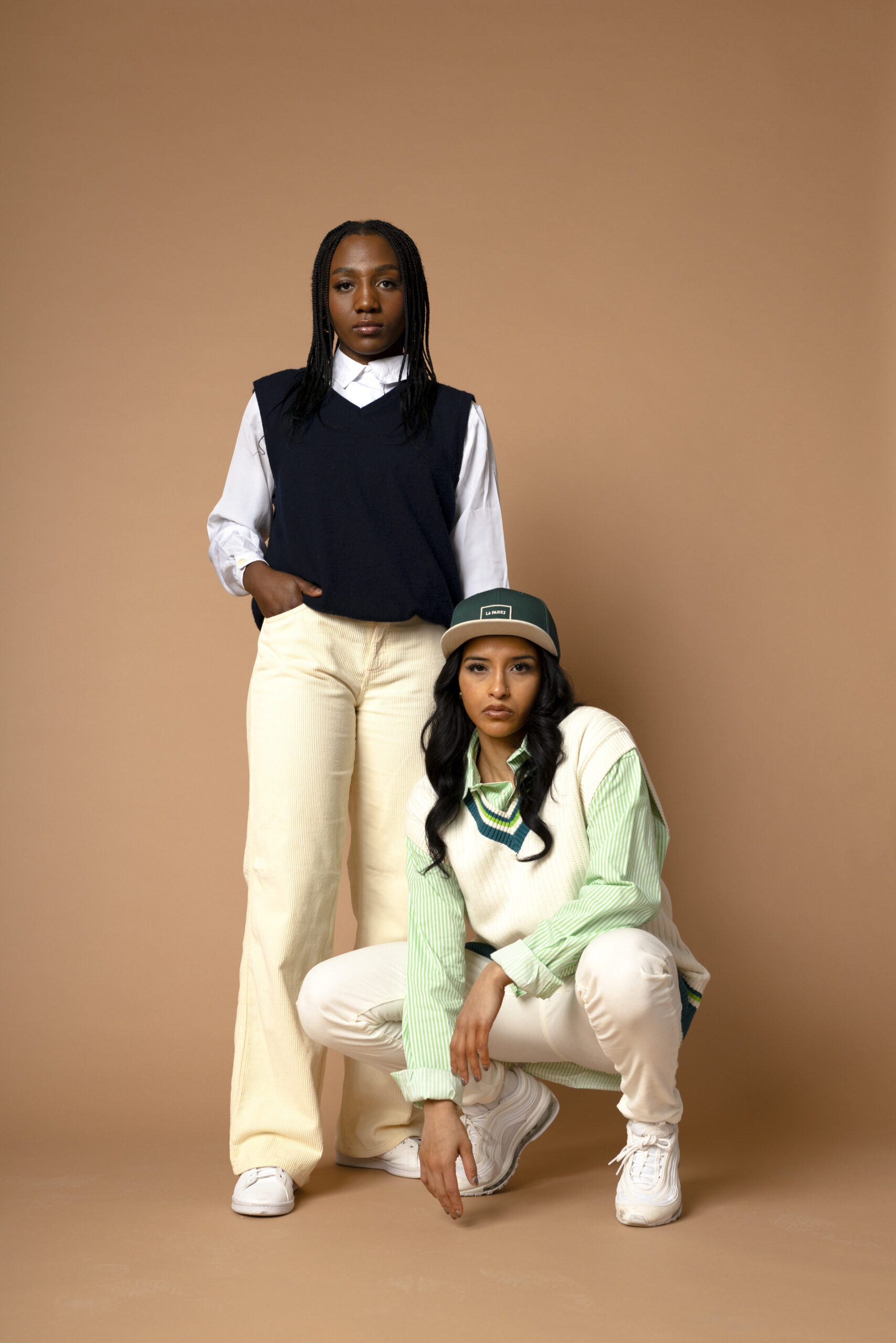 One model standing with one hand on a chair and one model sitting in the chair with their hand resting under their chin while wearing retro sports clothing against a brown backdrop.