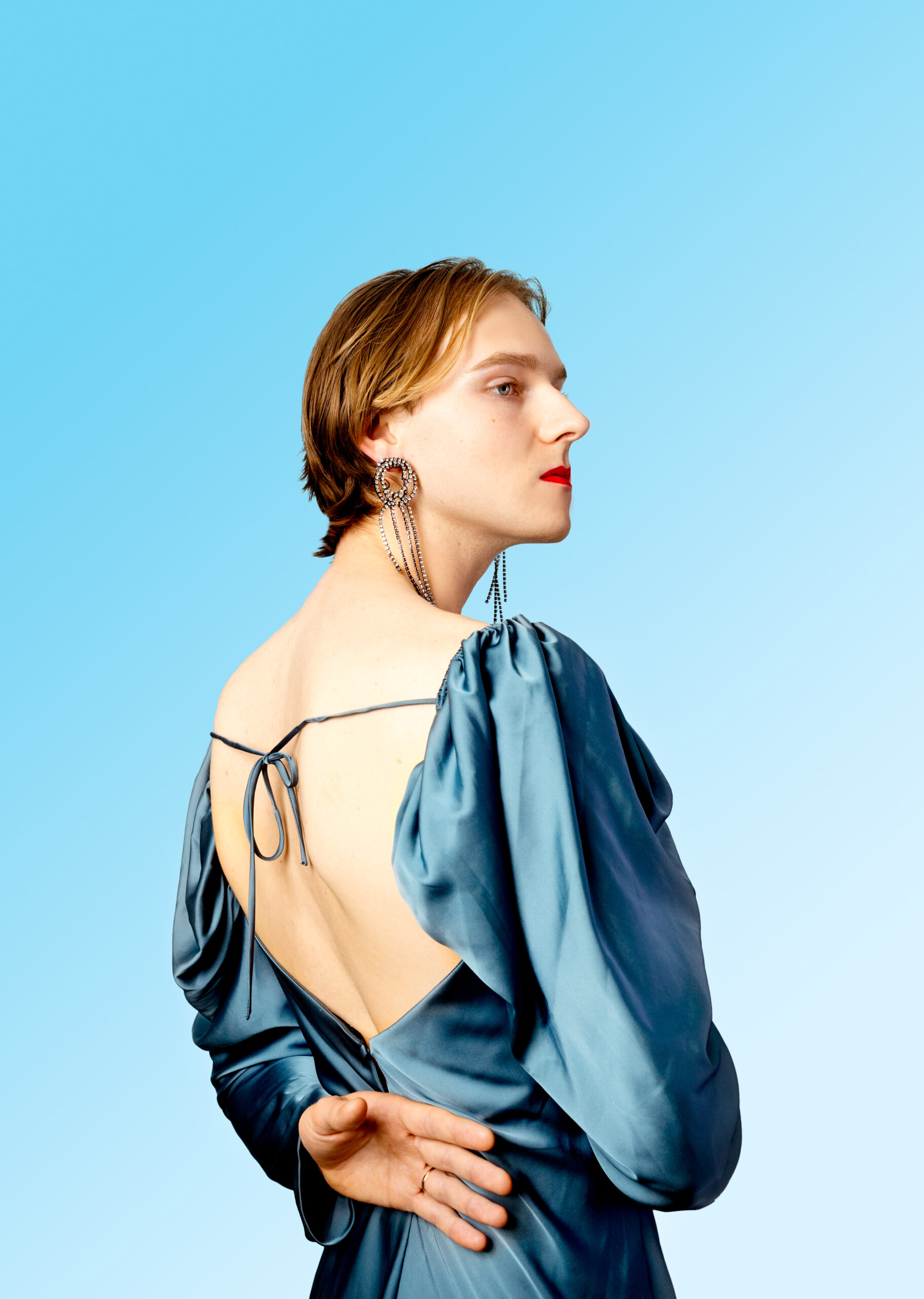 White model standing with their back at an angle with one hand resting on their lower back while wearing a blue silk dress, red lipstick, and silver earrings against a blue gradient backdrop.