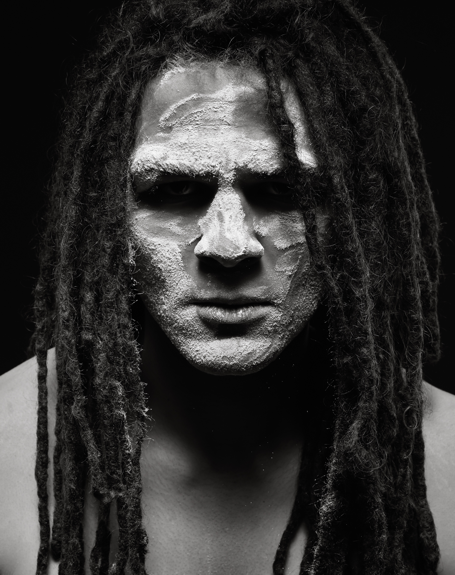 A man with long hair in dreadlocks is facing the camera head on, gazing into the lens. His face is covered with a white powder as some falls off his face the moment the photo was taken.