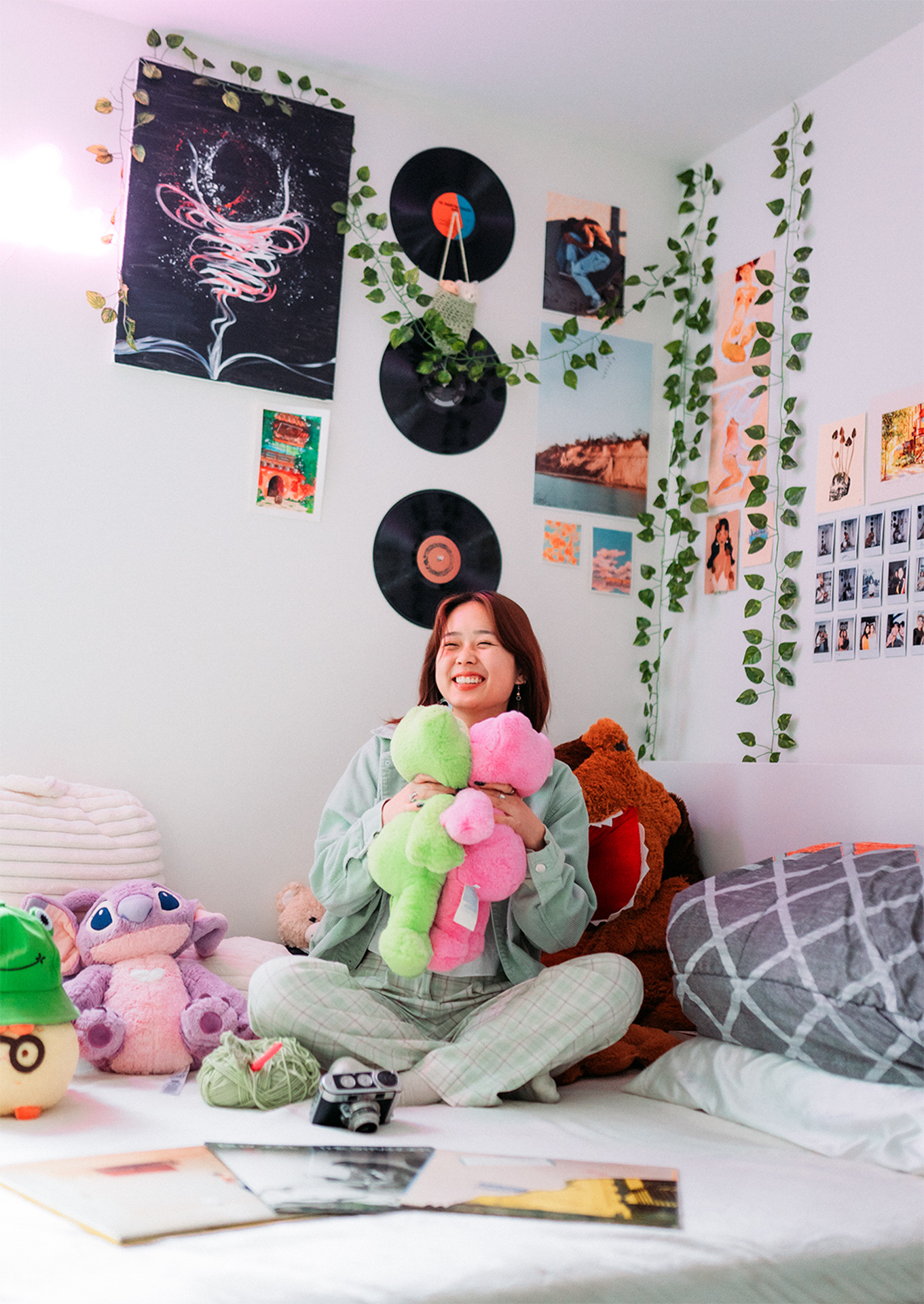 A portrait of a someone surrounded by stuffed animals and various objects of their room, they sit on their bed and smile while holding two stuffed animals in both hands
