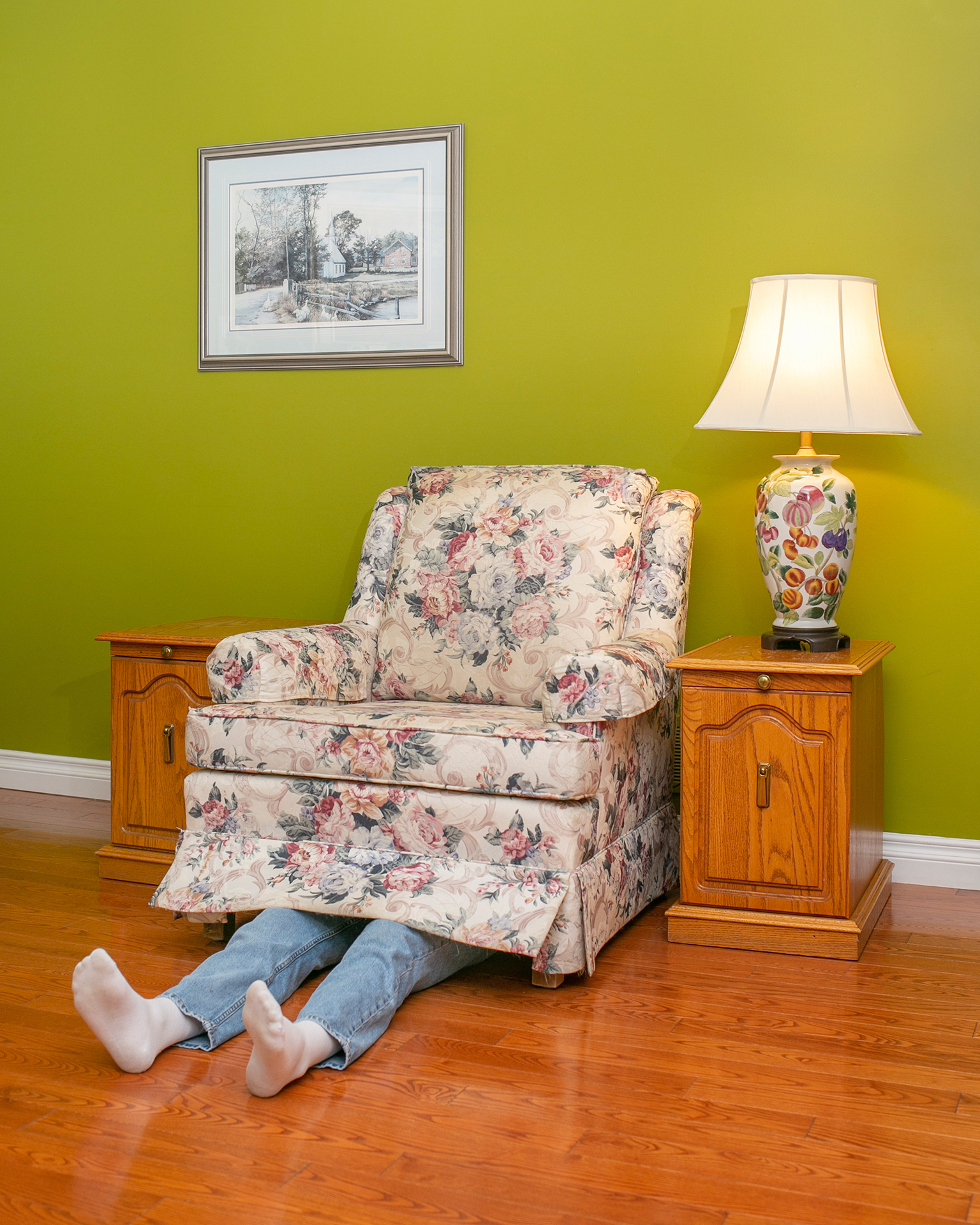 An empty white floral armchair placed against a green wall. There is a photo on hanging on the wall above the chair. Two small wooden side tables sit next to the chair with a lamp on one of the side tables. A set of legs wearing blue jeans and white socks are sticking out from under the armchair on the floor in a relaxed manner.