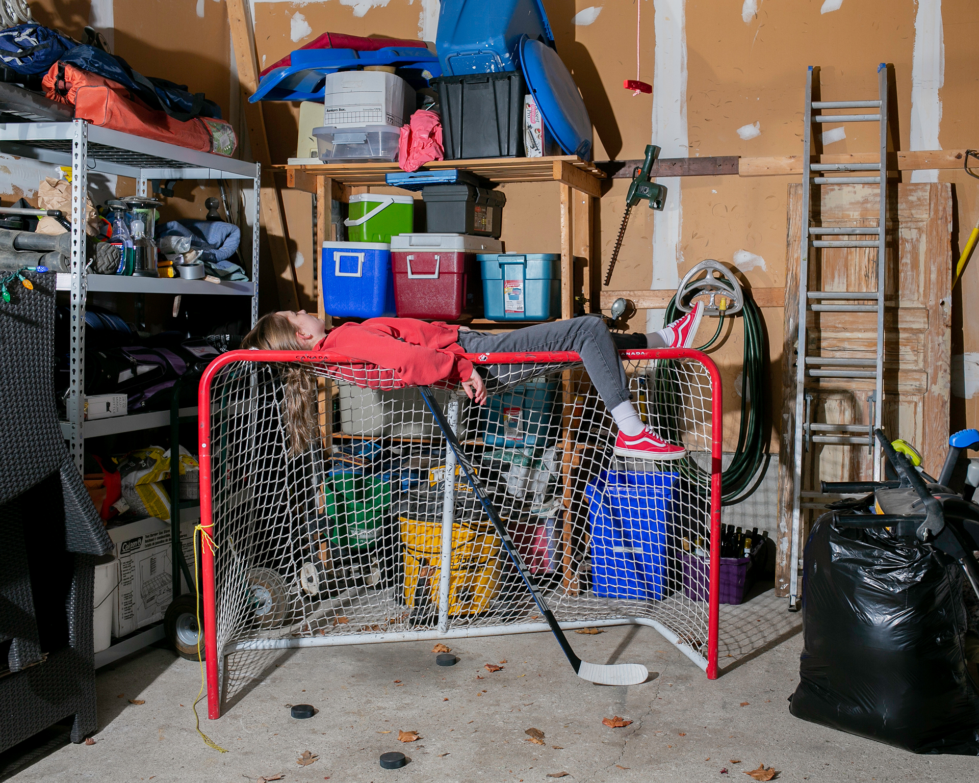 A person lies on top of a hockey net in a garage space. There is a storage shelf with various items on it behind the net. There are some pucks and leaves spread out on the ground.