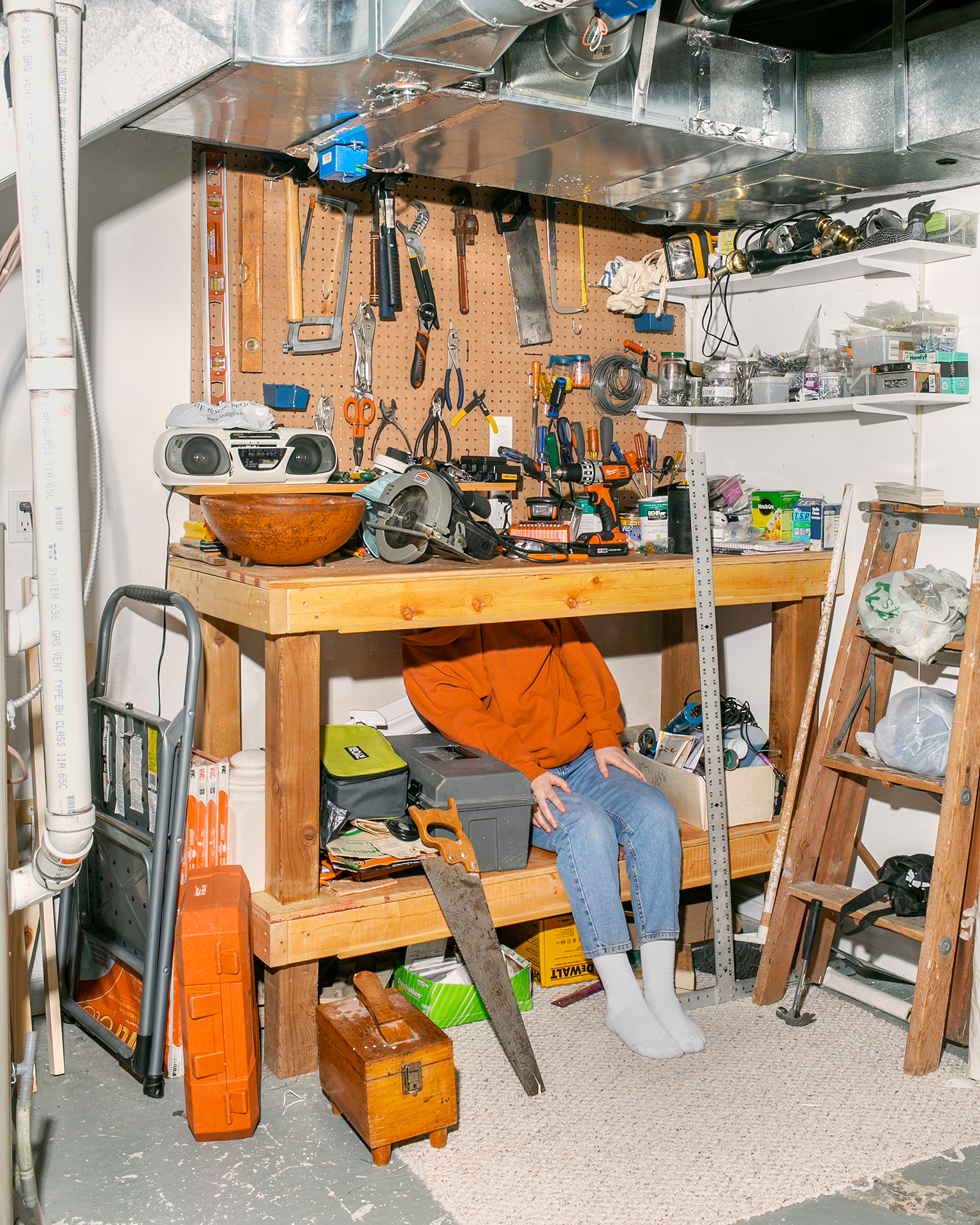 A person sits on a shelf of a wooden workbench, awkwardly hiding their face from view. There are a various tools hanging on the wall about the bench, as well as spread out on the surface of the bench. There is a saw leaning against the shelf on the floor. A metallic vent runs through the top of the frame.