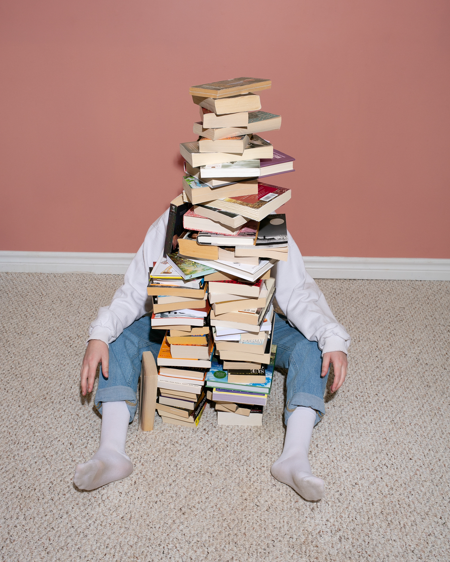 A person sits behind a tall stack of books with their legs sticking out from either side. The books completely hide the face of the person in the photo.