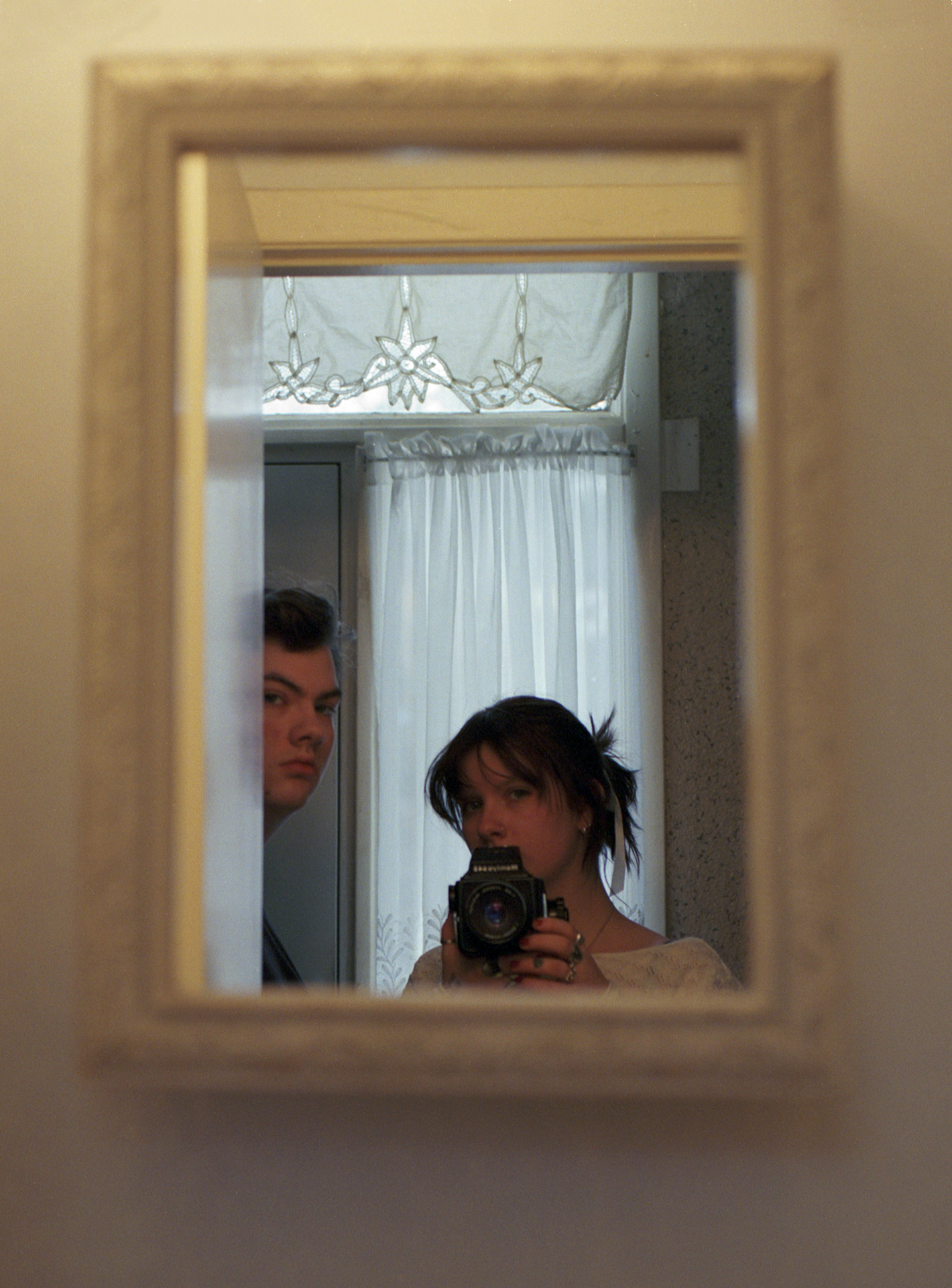 A brother and sister in a reflection of a mirror, they both confront the camera, but the brother is almost out of frame.