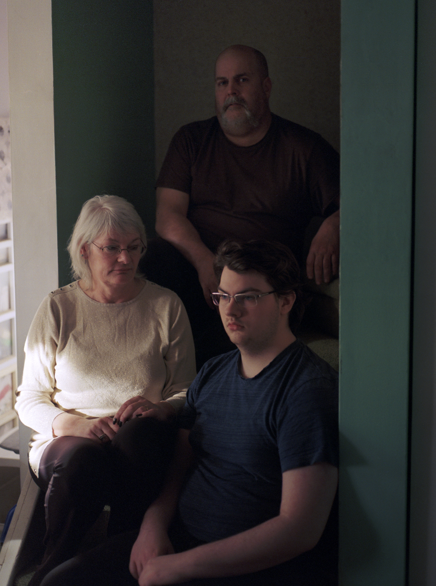 A take on a family portrait as they all sit posed on a staircase, a mother sits hand on lap staring at her son, her son looks off into the distance, and the father confronts the camera.