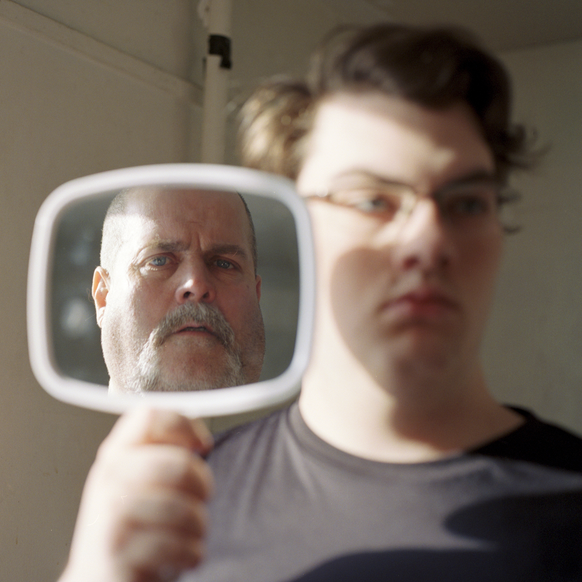 A man and son, the son holds a mirror that reflects the image of his father's face.