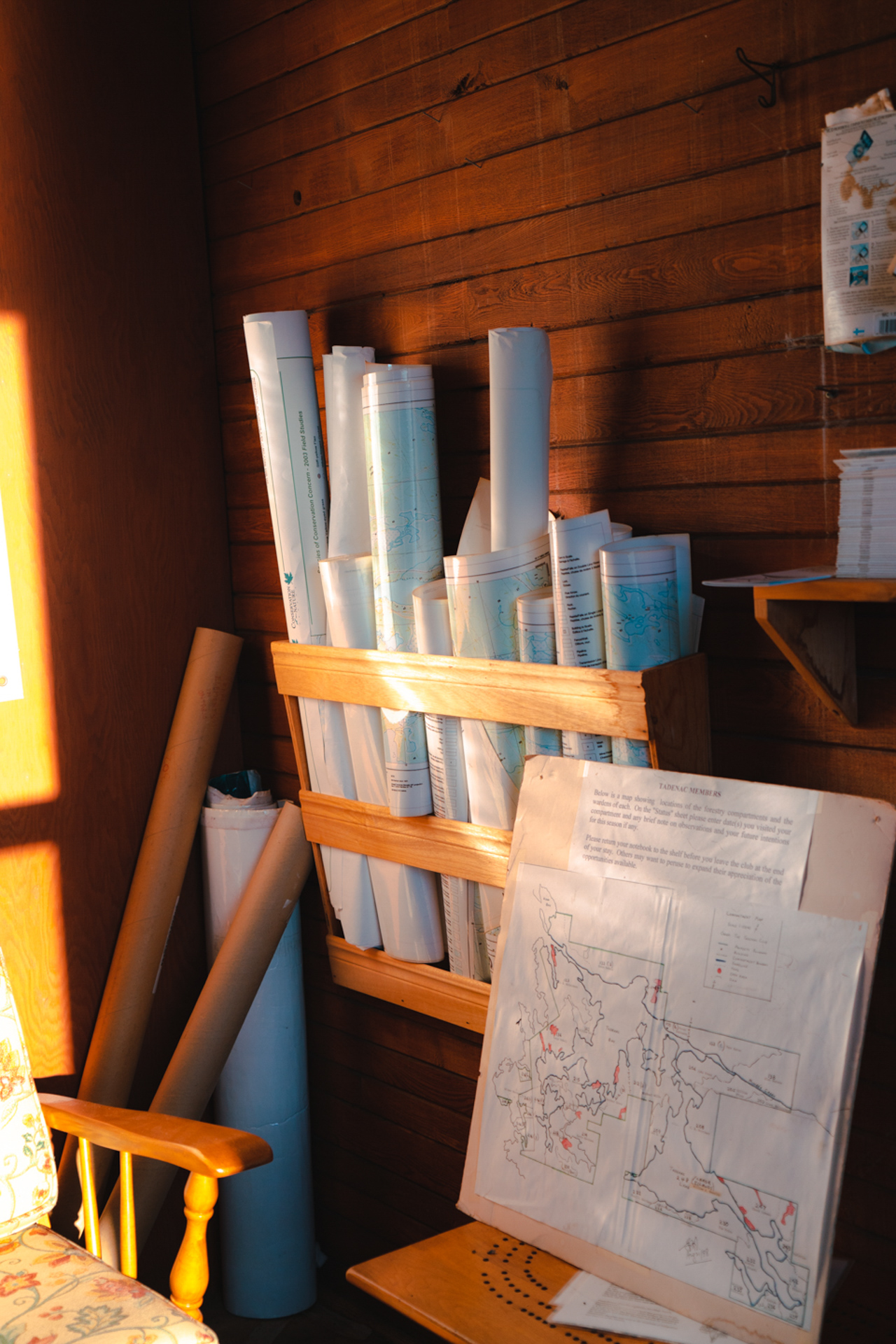 This photo consists of a brightly lit wooden cabin room. There are several maps rolled up in the corner of the room and stored on a hanging shelf. There is also a map on a wooden stand with colored outlines and descriptions.