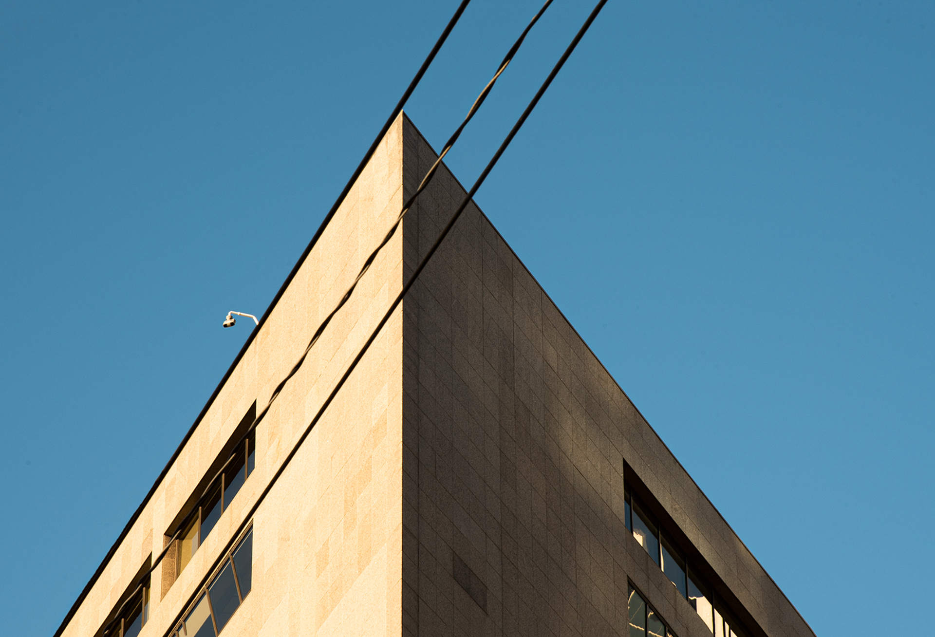 A photo looking up at a corner of a building, three power lines align with the corner of the building diagonally across the image. The sky is a bright blue that matches well with the beige exterior of the building.
