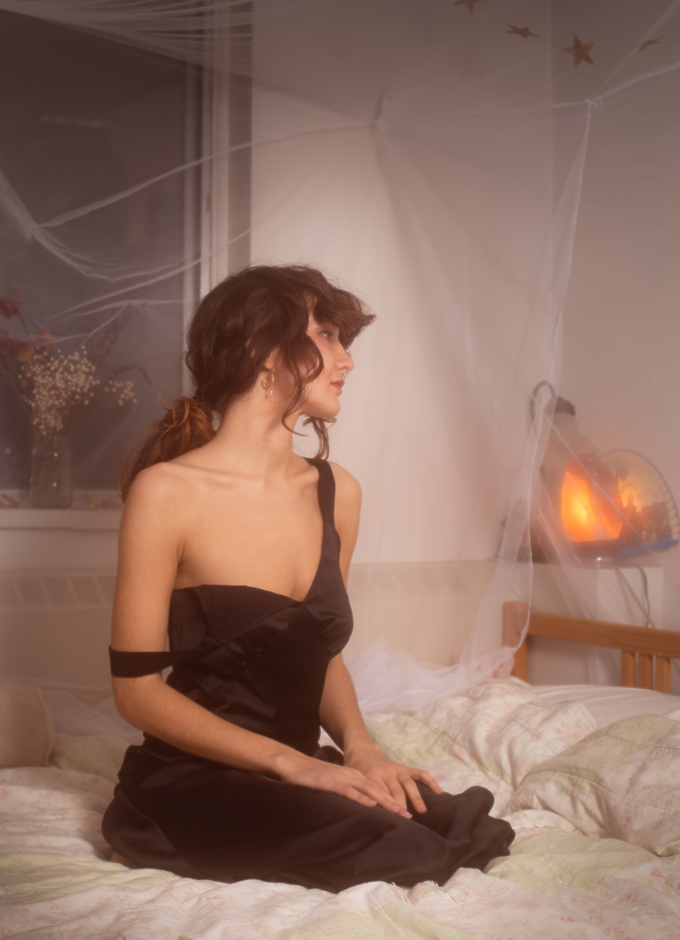 Lola kneeling on their bed. Their head is turned to the side, away from the camera showing their profile. They are wearing an elegant black silk dress with thick straps, one strap has fallen off their shoulder. In the background of the image there are some dried flowers in a vase on their windowsill behind them, and a salt lamp is sitting behind their headboard to the right. Their hands are placed elegrantly on their lap.