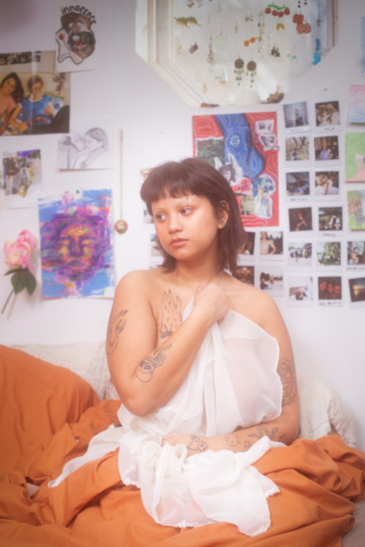 Maya sitting on her bed with orange sheets at the base of her body and a white silk sheet covering the front of her naked body. The walls of her room are covered in her artwork and pictures she took. She is looking off to the side.
