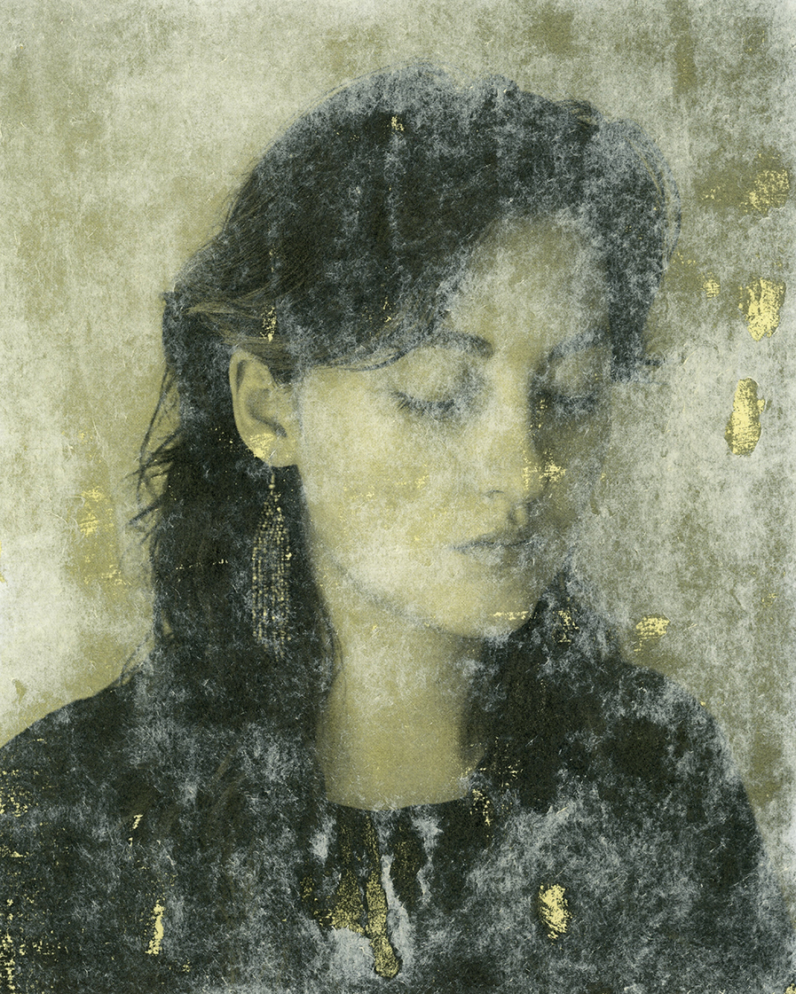 Self Portrait: A black and white portrait of the artist against a plain backdrop, who is turned 3/4, facing the left and looking down towards the ground. They are wearing a pair of fringe beaded earrings and a black shirt. The image has been transferred to a piece of watercolour paper dyed with Sweetgrass that is light green in colour.