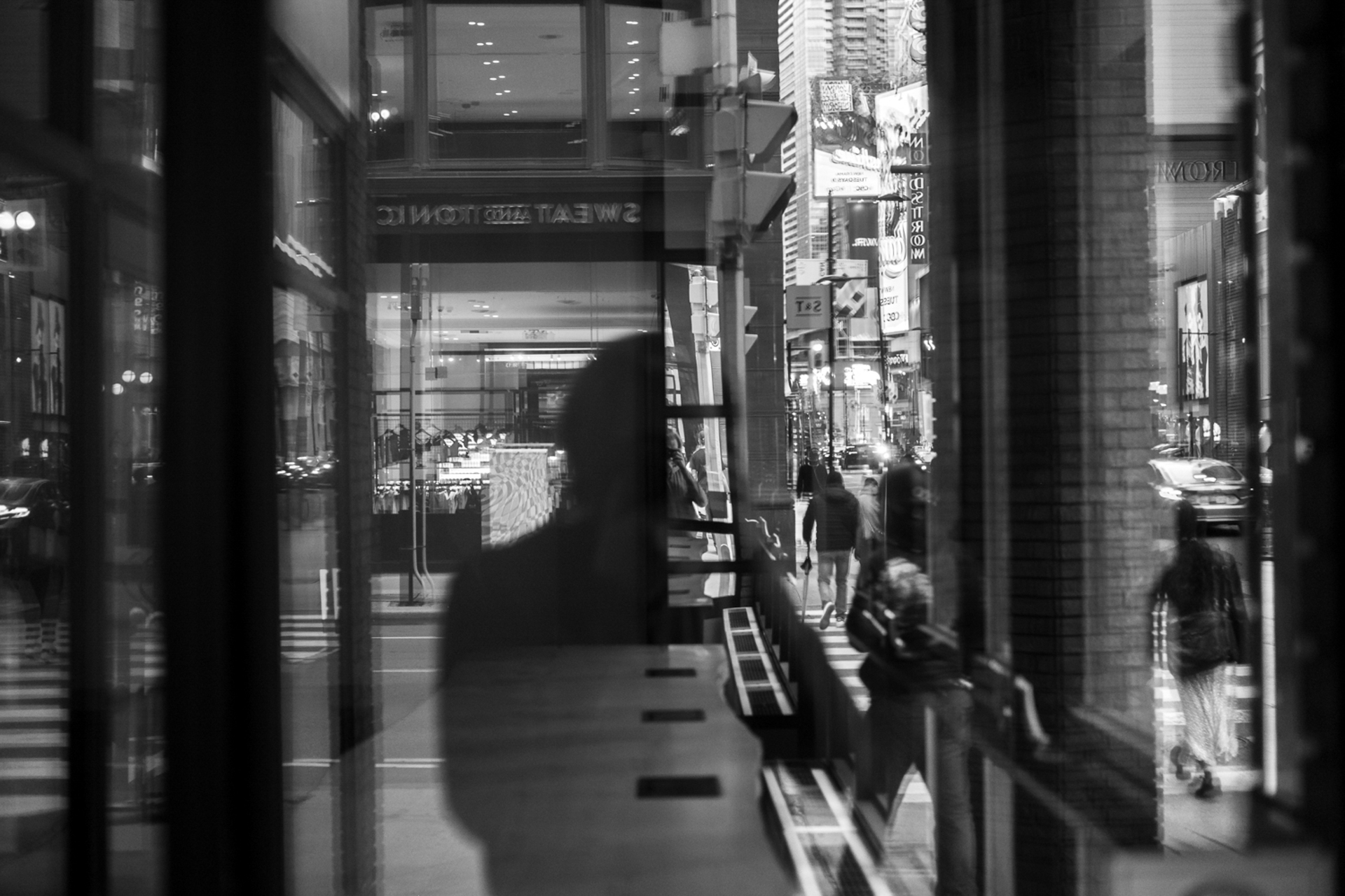 This photo consists of the reflection of a man’s silhouette and the streetscape with several moving people and cars on the right side of the photo.
