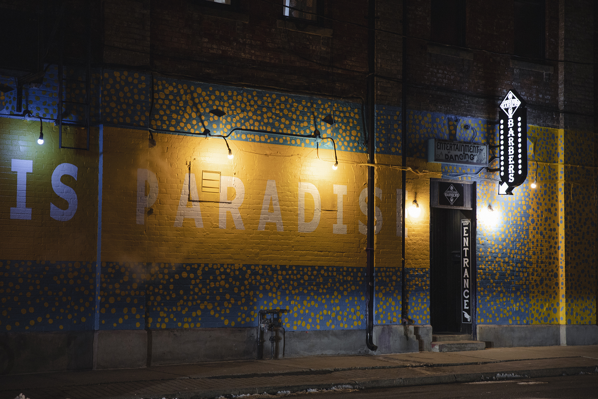 An empty sidewalk and building wall at night. The wall has some unpainted bricks on top, but most of it is painted with a blue and yellow speckled design. In the middle of the wall is a yellow banner that has the words "IS PARADISE" painted across it. After these words, there is a small black door that says "entrance" on it, as well as a glowing sign with an arrow pointing towards the door.