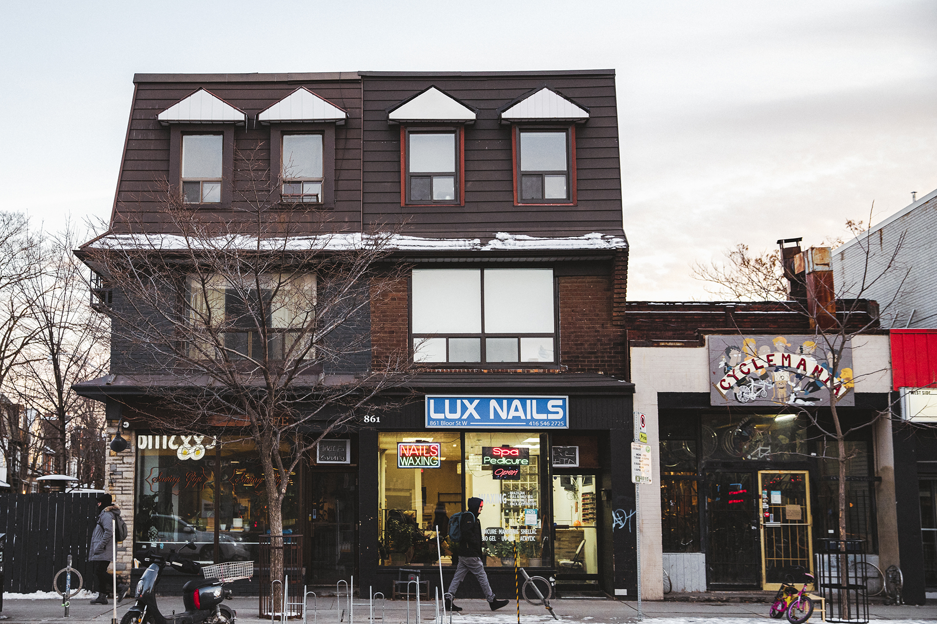 A small townhouse-style building photographed from across the street. There is a sign on the building white says "LUX NAILS," indicating the building houses a nail salon business. In front of the building, there is a man walking by as well as a parked motorcycle. The light is soft and it appears as though it is sunset.