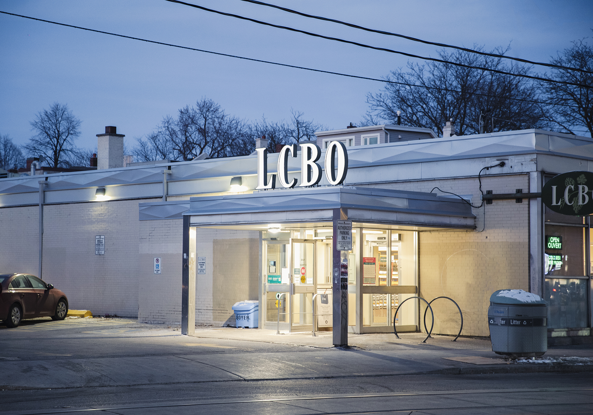 An all-white LCBO building photographed from across the street. The sun has started to set and there is little daylight left, so the light from the store and the LCBO sign makes it seem like it's glowing. There is one red parked car on the left side of the frame, and the tops of trees are poking out from behind the LCBO.