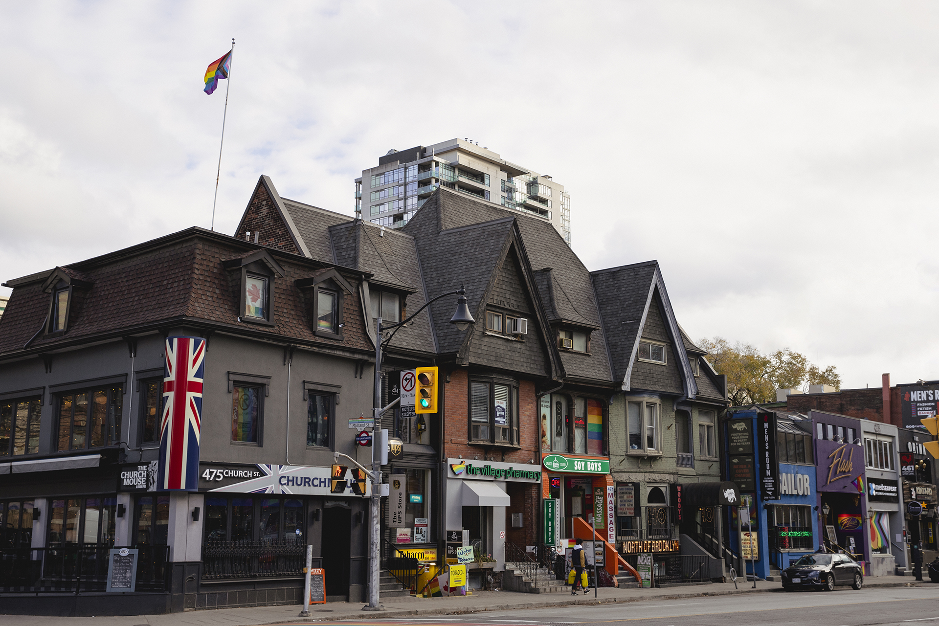 : A series of townhouse-style buildings photographed from across the road. There are numerous colourful signs strewn across the buildings, as well as an LGBTQIA+ flag being flown. The sidewalk in front of the buildings in empty with the exception of one person and one parked card.