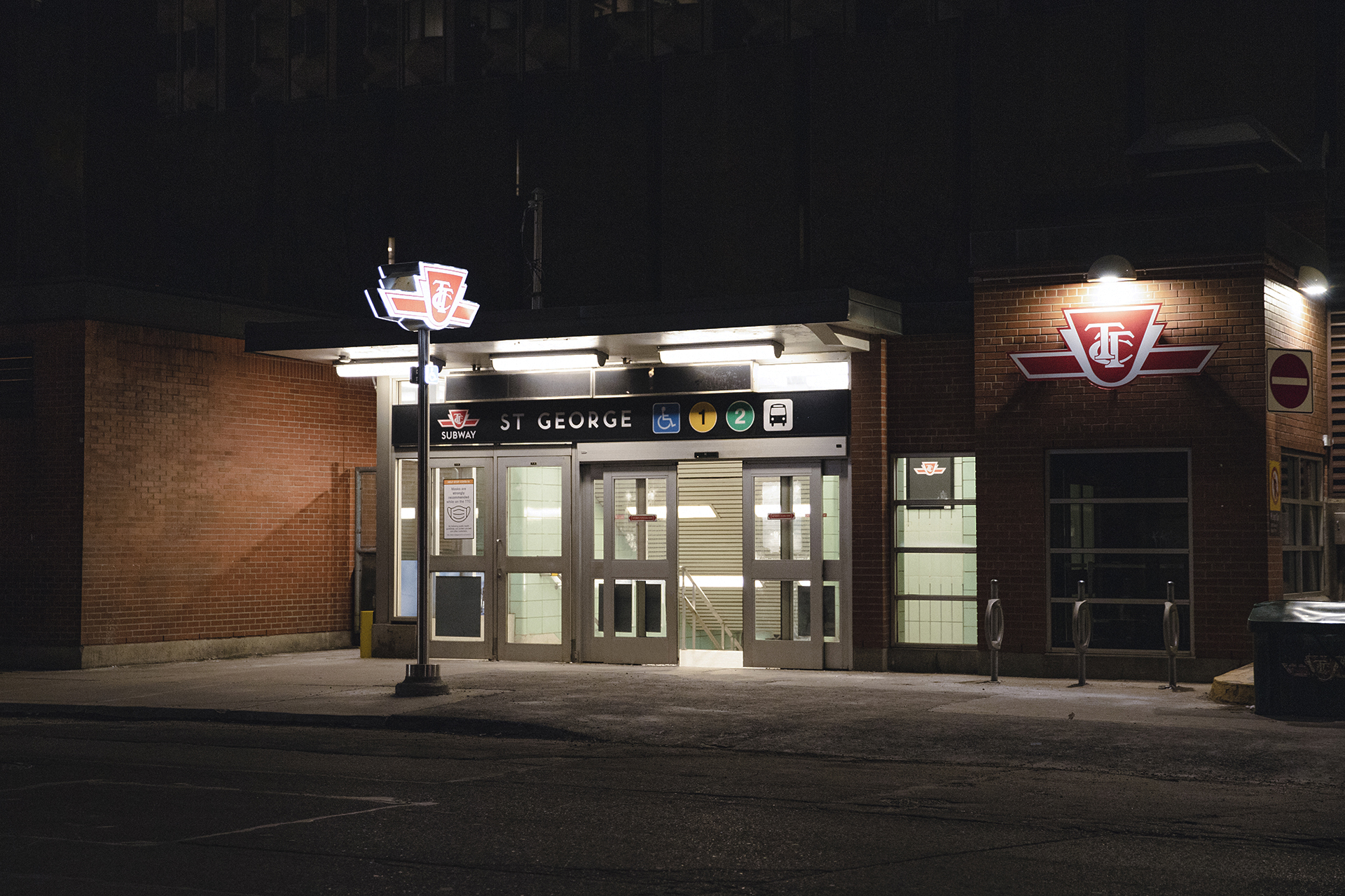 The St. George Subway station photographed from across the street. It is late at night and there are no lights coming from buildings in the background. The only light source in the image is from inside the subway station as well as a TTC sign outside on the sidewalk. The doors to the subway station are partially open but there are no people or cars.