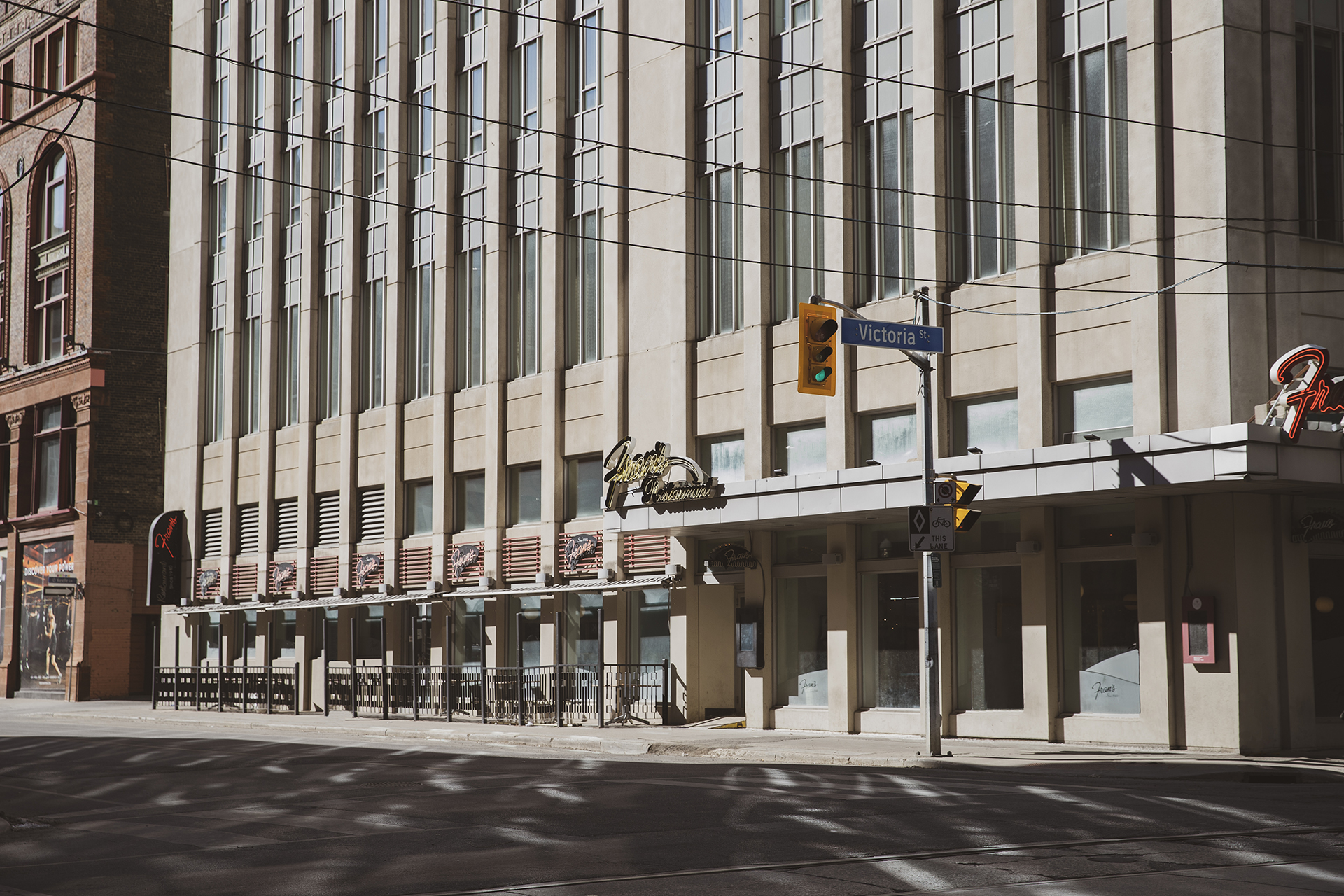 A tan-coloured building being bathed in sunlight. The building has many columns of tall windows and a golden sign that says "Fran's" on the side. There is a street sign and a stoplight, but there are no cars on the road. There is light from the building reflecting onto the pavement.