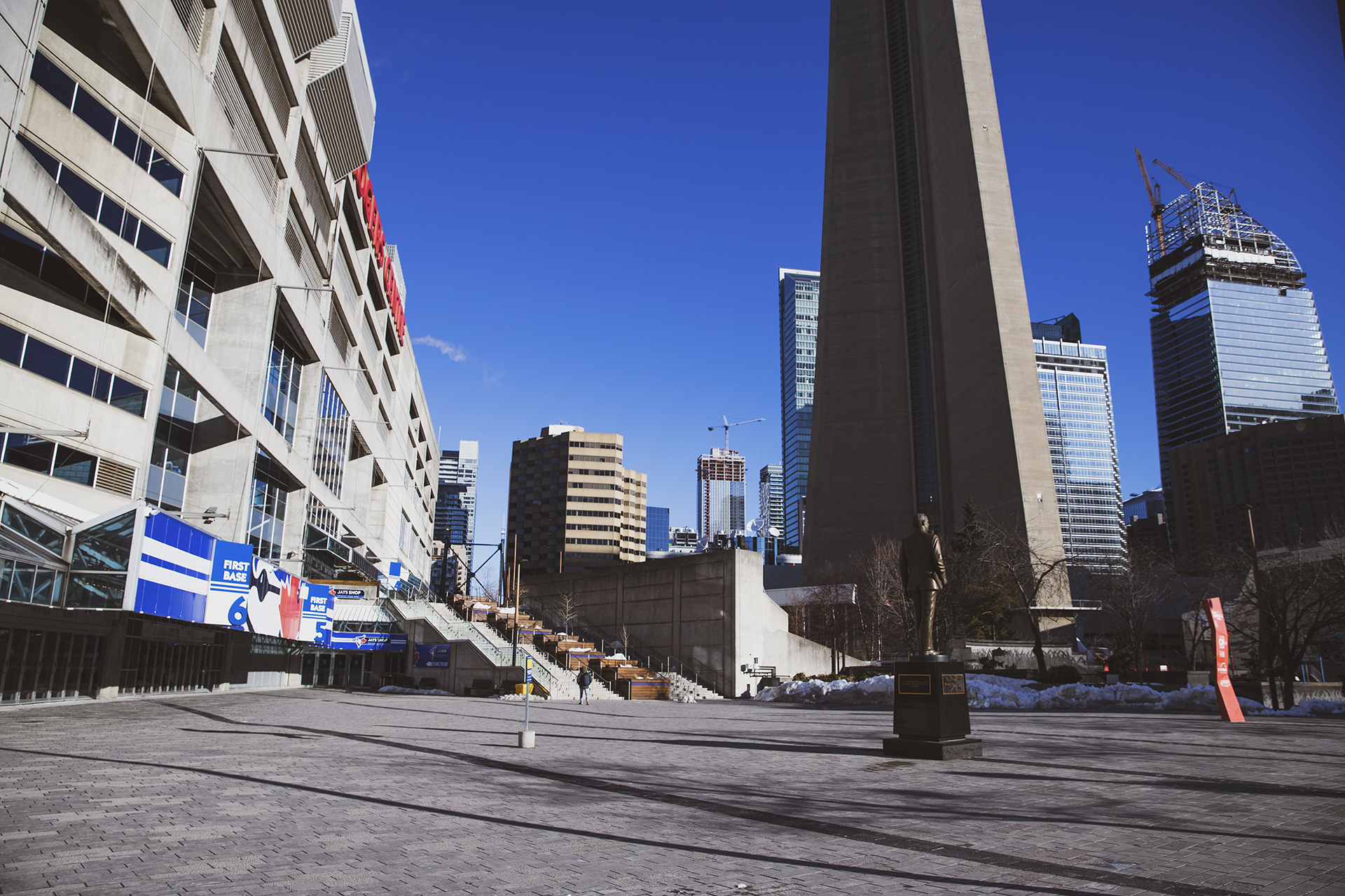 An empty sidewalk outside of the Rogers Centre. The Rogers Centre is on the left, with a bronze statue of a man on the right. The base of the CN tower can be seen behind the statue, as well as some buildings in the distance.