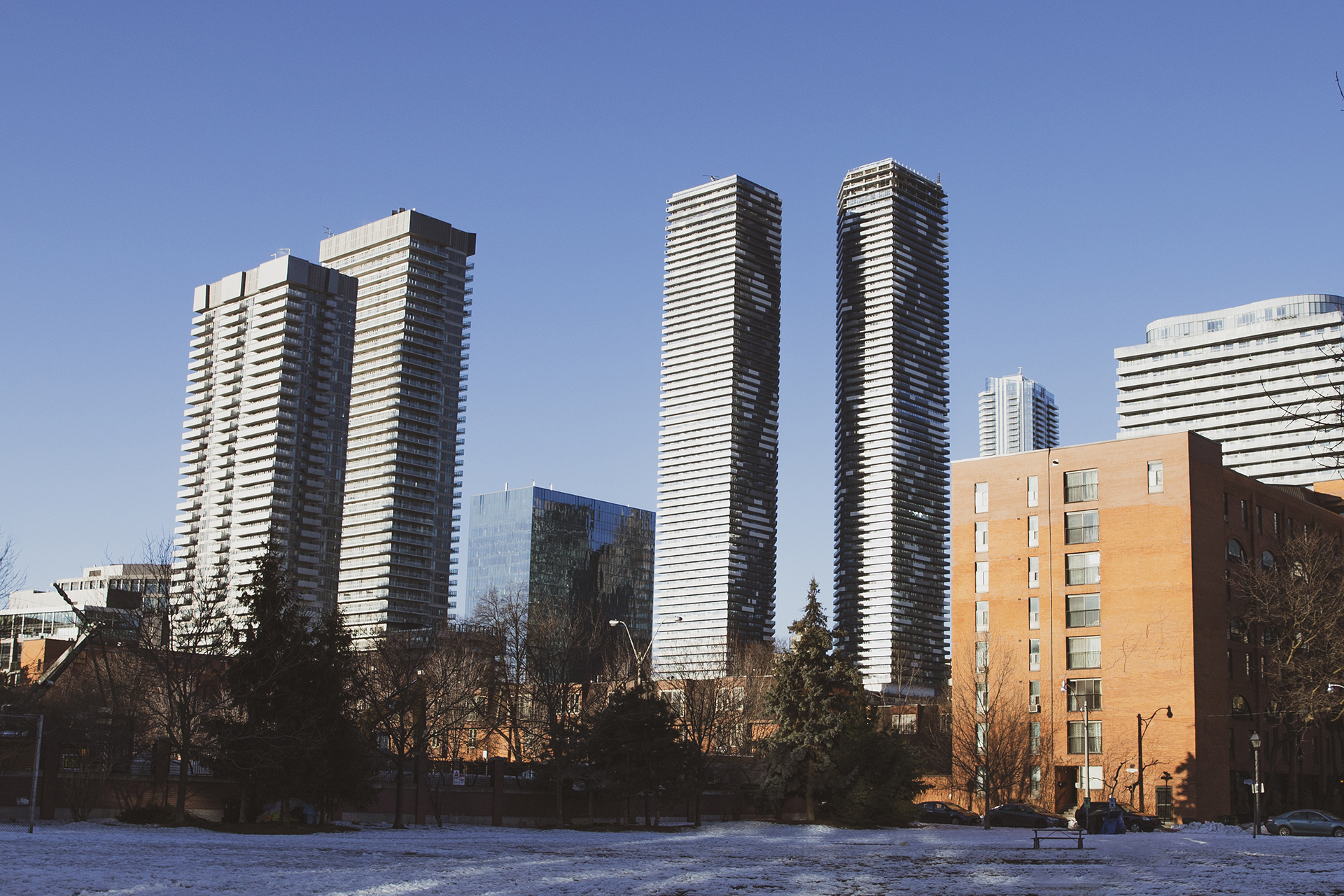 A snow-covered park in the early morning. There are a few trees scattered across the background. There is one 8-story, brick apartment building, and multiple tall glass buildings in the distance.