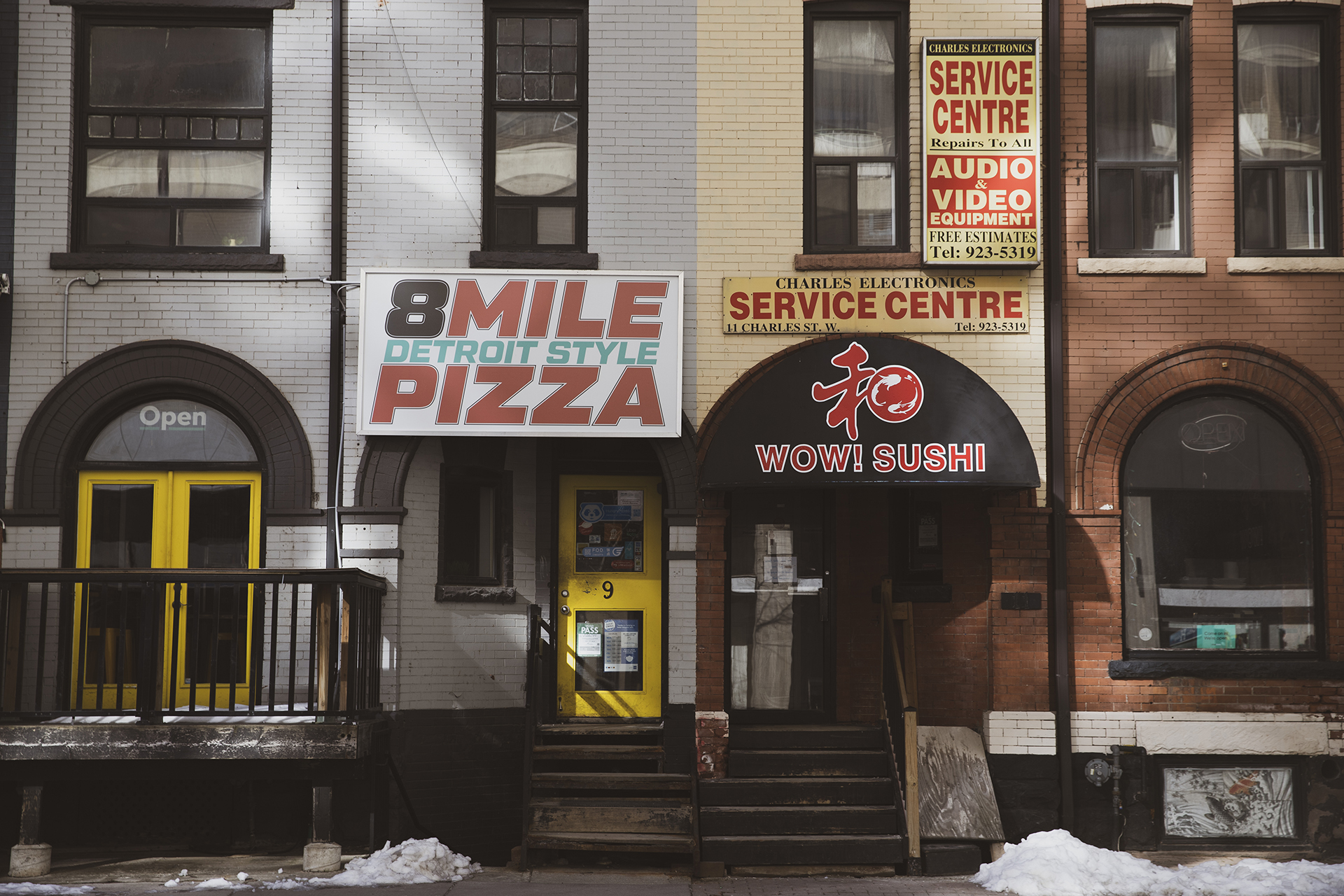 Two businesses side by side: a pizza restaurant and an audio and video equipment store. The pizza business is painted grey with yellow doors, and the equipment store has pale yellow and red bricks.