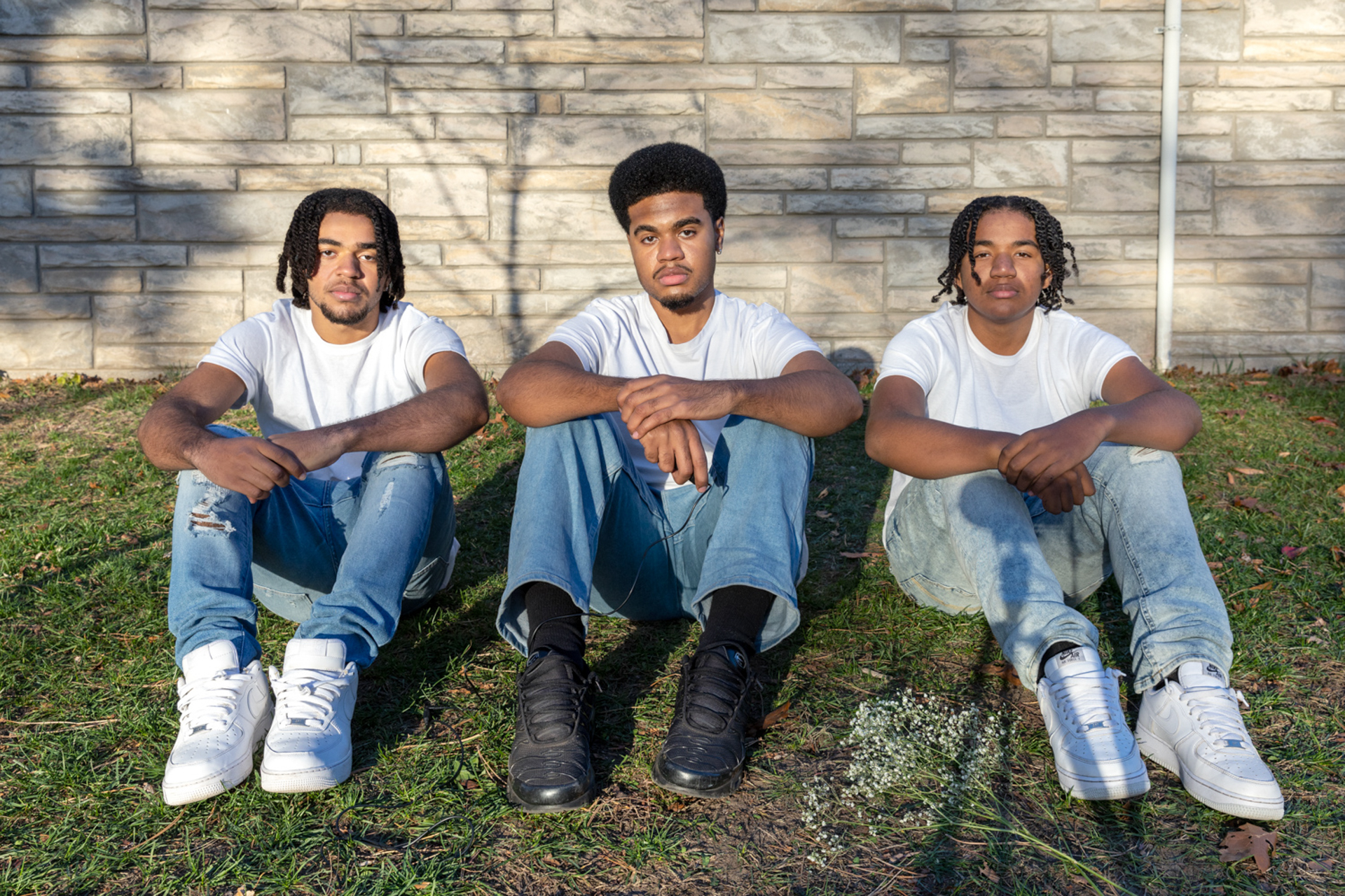 This photo consists of three Black men who are sitting in a grass field on a bright day, all gazing into the camera. They are all wearing white short-sleeved shirts and light-blue jeans. They wear alternating color shoes; white, black, white. The same goes for their hair, the first is twisted, the second is an afro and the last is twisted as well. The three of their arms are crossed over their knees, the man in the middle holds a black shutter release cable and a bunch of Baby’s Breath flowers lay to the right of him.