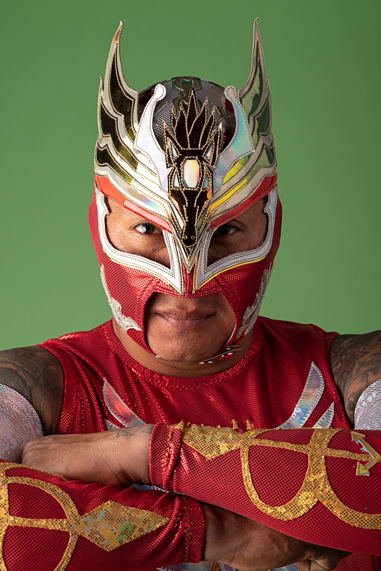 This photo is a portrait of a Lucha Libre wrestler. The wrestler is wearing a red, gold, and silver mask with a pegasus and flared wings on the top head section. His arms are crossed displaying the gold and silver embroidery on the arm sleeves and t-shirt portion. The Lucha Libre is standing in front of a bright green background.