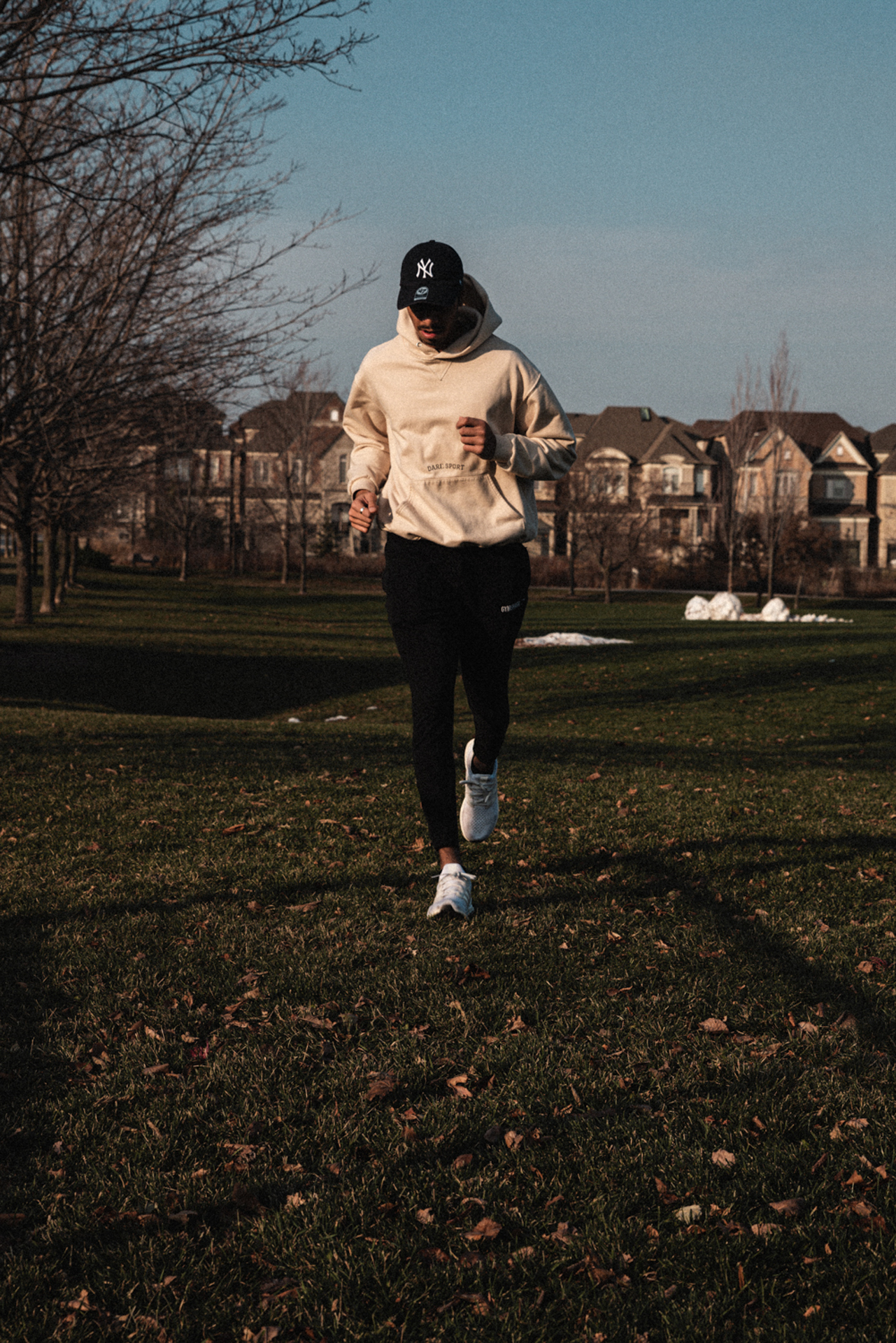 This photo depicts a man wearing athletic clothing running through a field on a bright day. There are houses behind him and brown leaves that cover the grass he is running on.