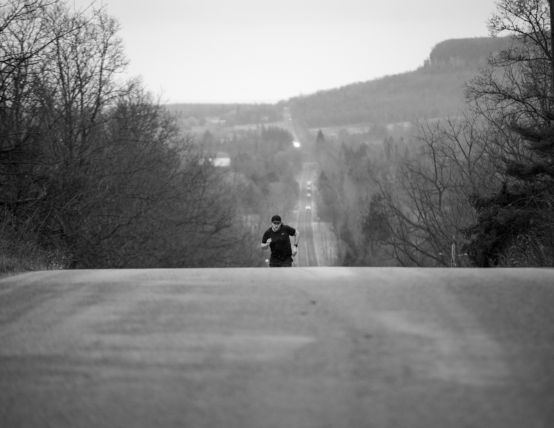 The photo depicted is in black and white. A man is running up a steep road. He is wearing all black and is captured striding up the road free of cars.