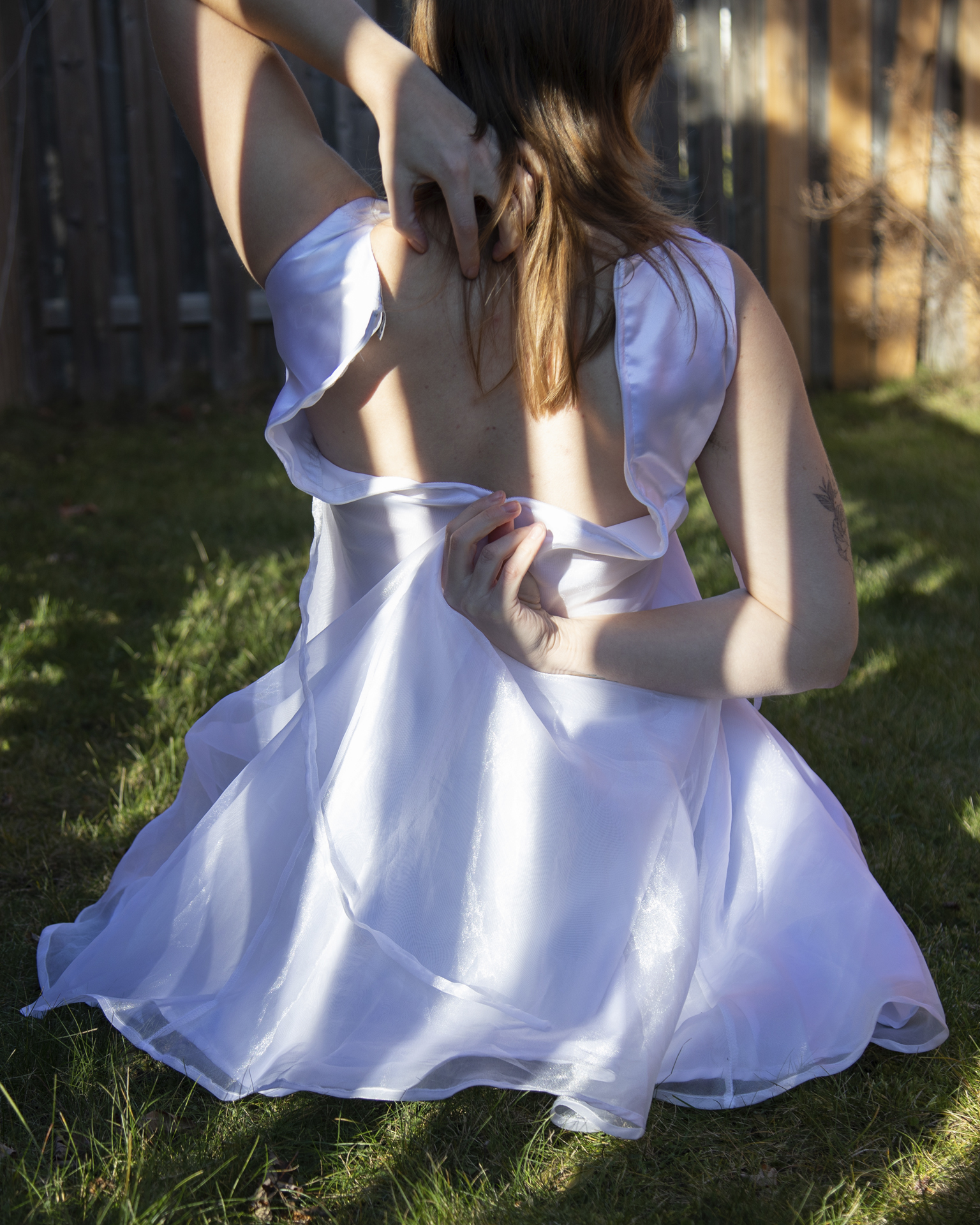 But it just won't fit: A woman kneels on grass while facing away from the camera, she is trying to zip up a white communion dress that is too small for her.